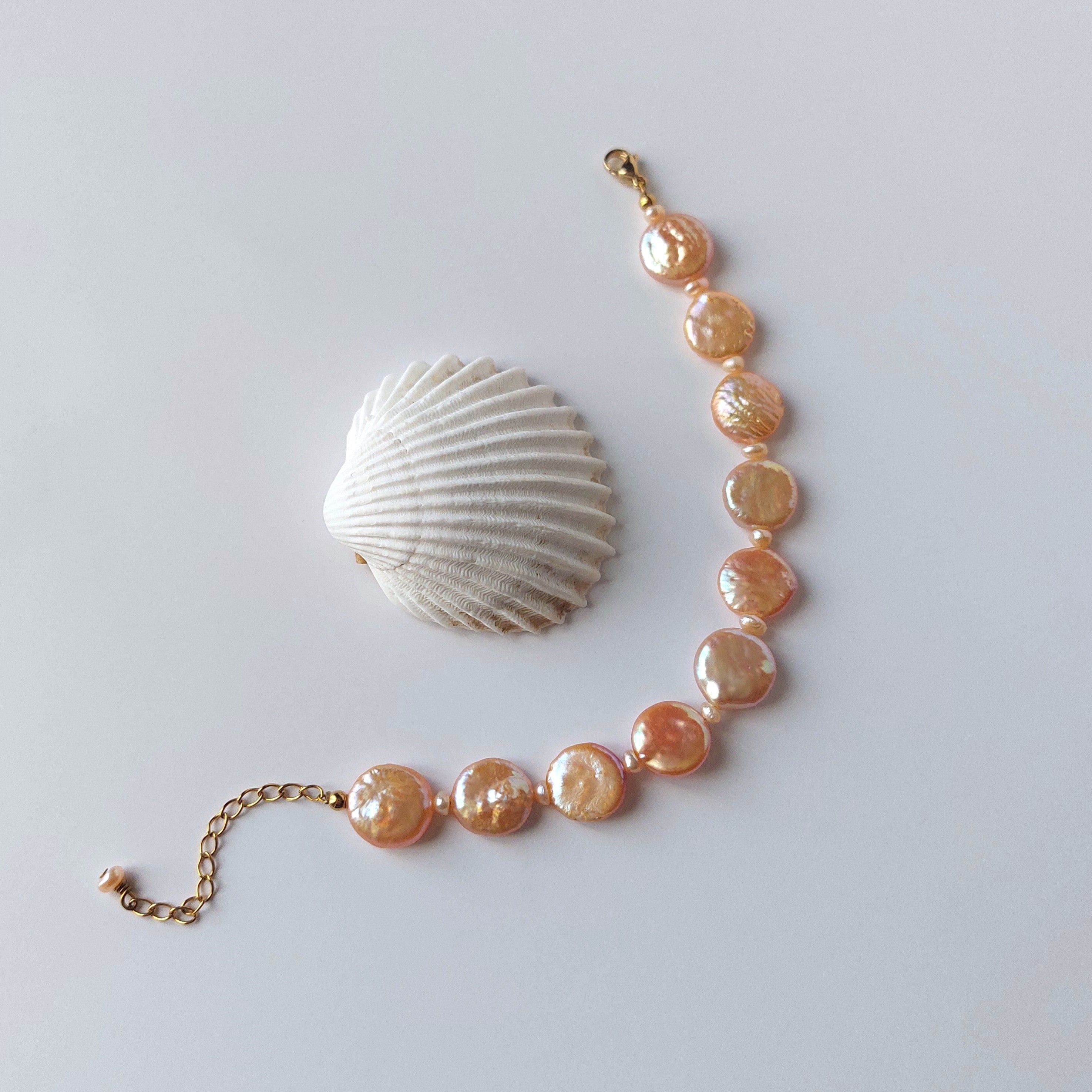 Bracelet en acier inoxydable doré avec perles d’eau douce naturelles oranges plates, bijou lumineux posé à côté d’un coquillage, sur fond neutre.