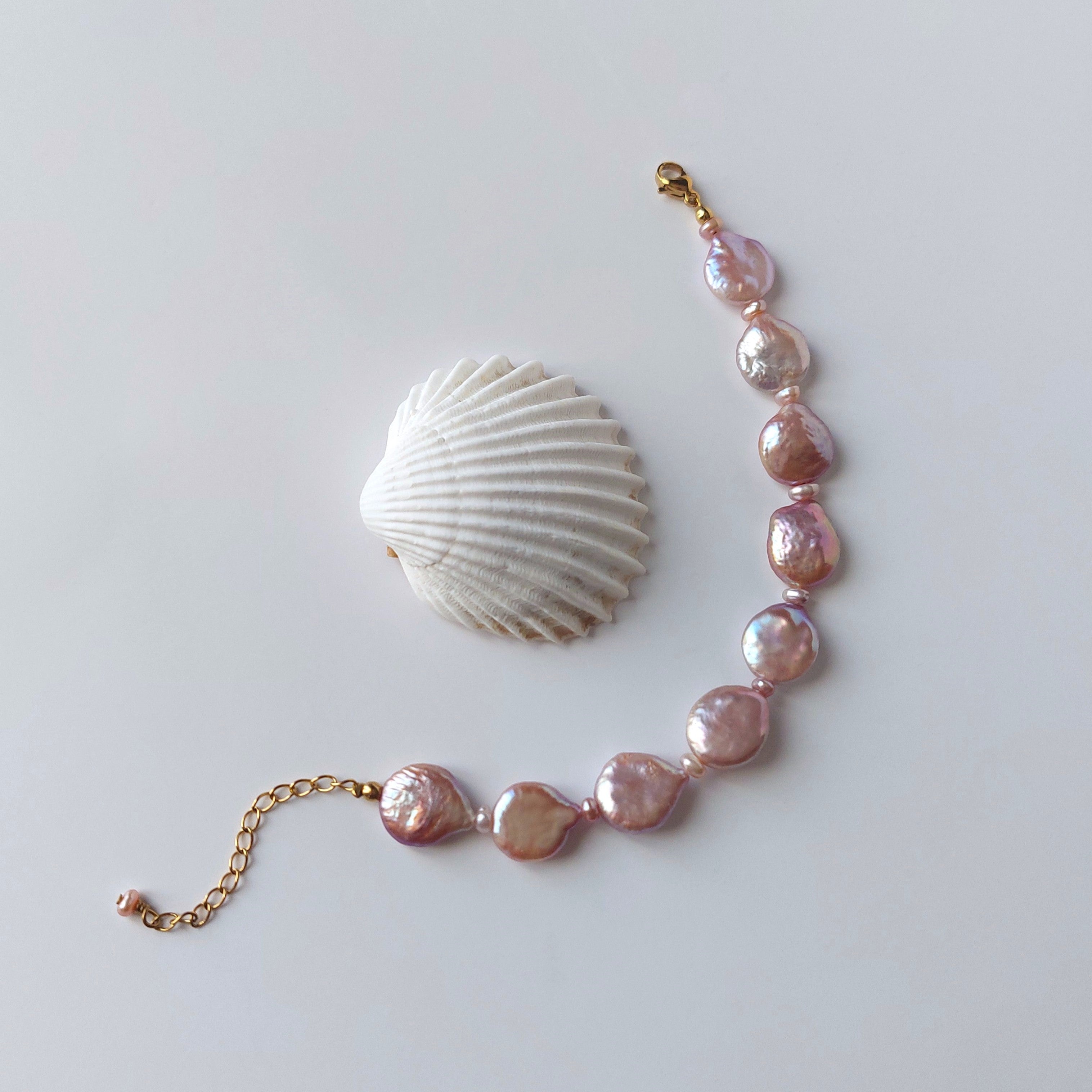 Bracelet en acier inoxydable doré avec perles d’eau douce naturelles violettes plates, bijou lumineux posé à côté d’un coquillage, sur fond neutre.