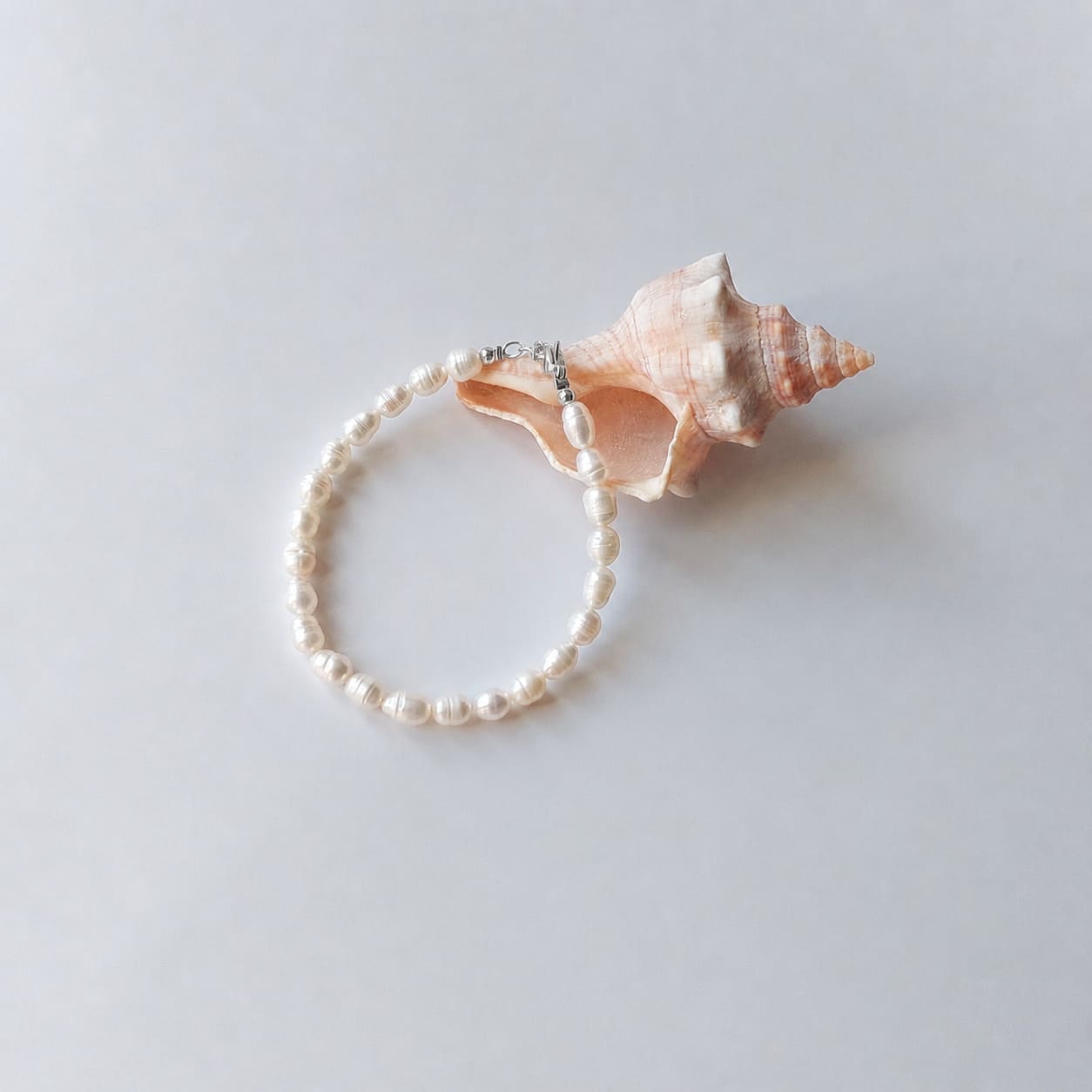 Bracelet entièrement composé de petites perles d'eau douce blanches ovales nacrées avec un fermoir argenté, disposé en cercle sur fond gris clair aux côtés d'un grand coquillage en spirale rose et beige.