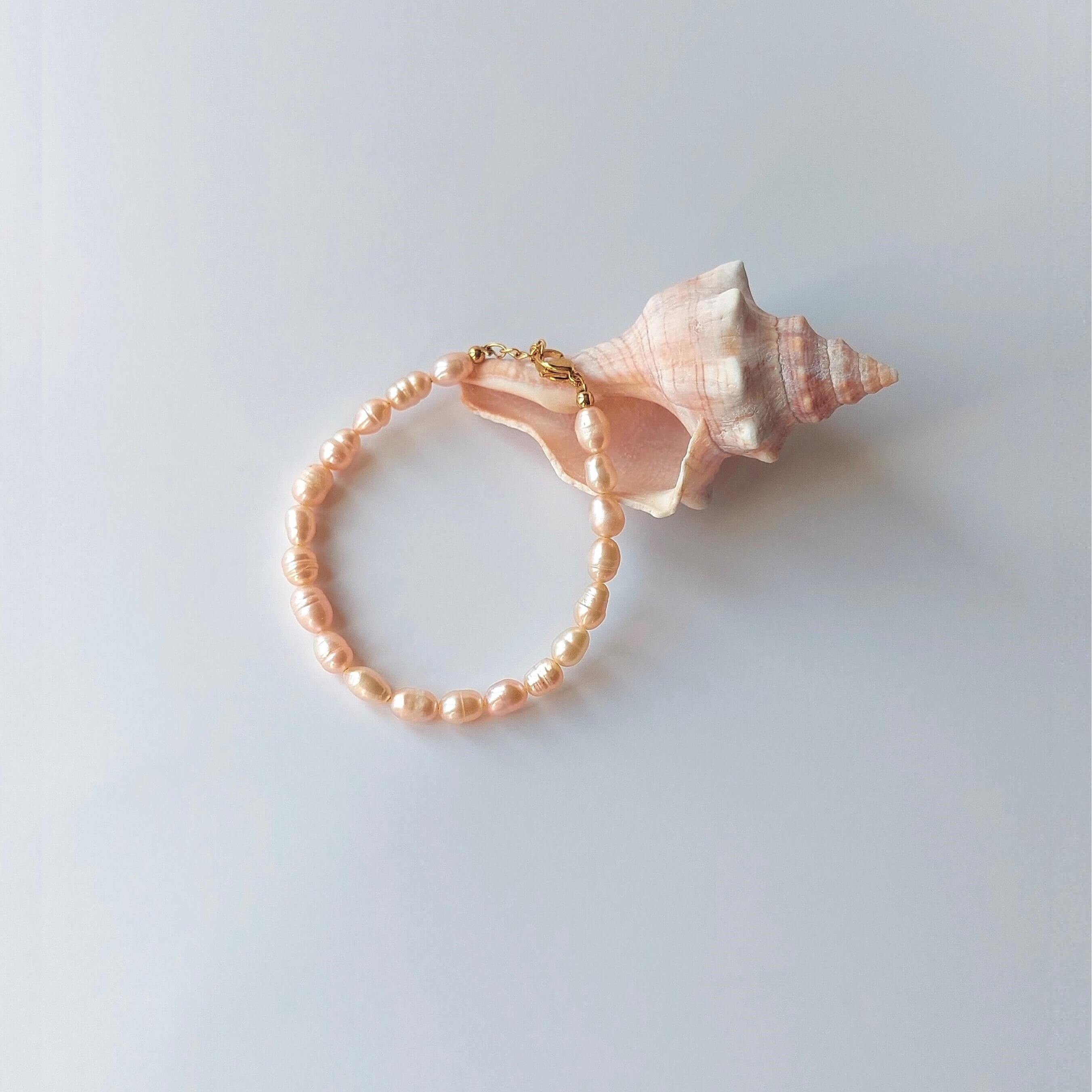 Bracelet en acier inoxydable doré orné de perles d’eau douce naturelles oranges, posé devant un coquillage rose, sur fond neutre.