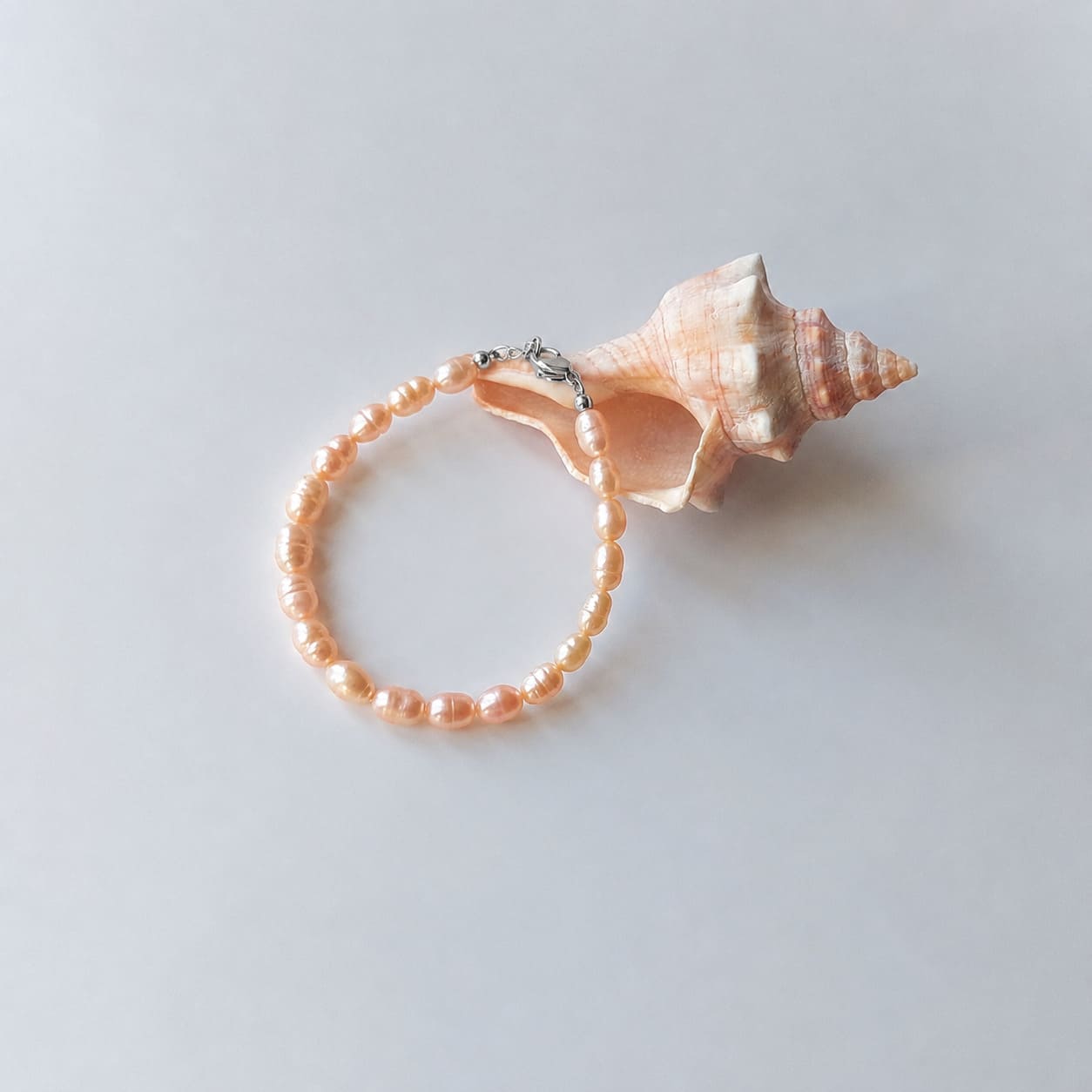 Bracelet entièrement composé de petites perles d'eau douce oranges ovales nacrées avec un fermoir argenté, disposé en cercle sur fond gris clair aux côtés d'un grand coquillage en spirale rose et beige.