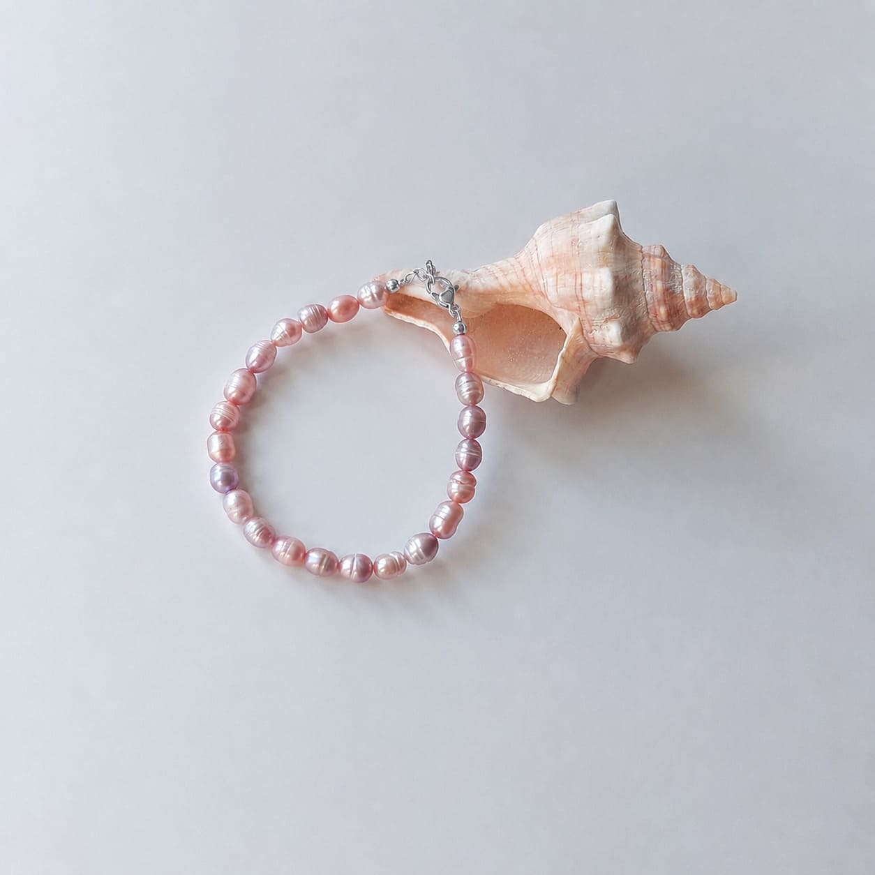 Bracelet entièrement composé de petites perles d'eau douce violettes ovales nacrées avec un fermoir argenté, disposé en cercle sur fond gris clair aux côtés d'un grand coquillage en spirale rose et beige.
