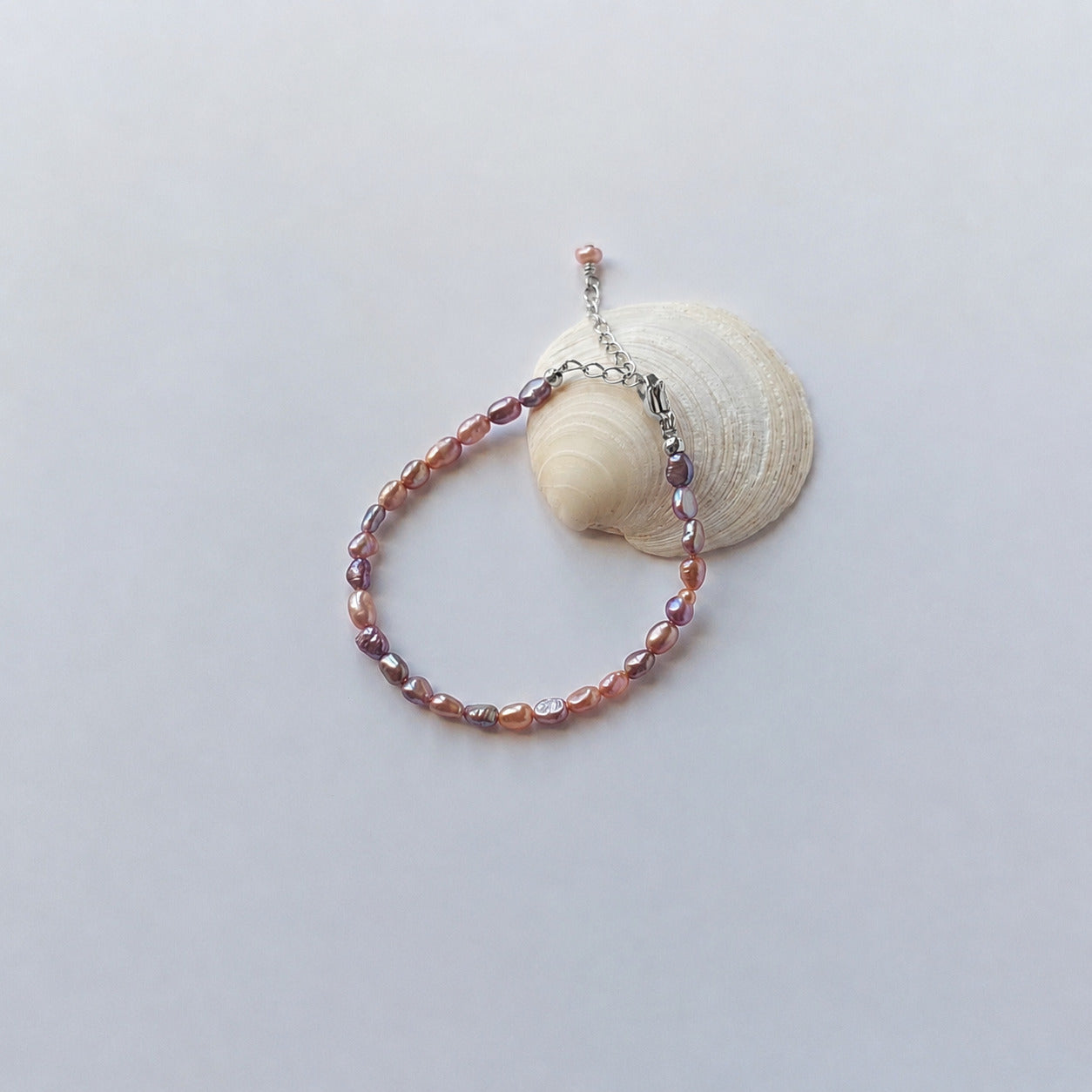 Bracelet entièrement composé de petites perles d'eau douce ovales de couleur violette nacrée avec un fermoir mousqueton argenté et une chaînette d'extension, disposé en cercle sur fond gris clair aux côtés d'un coquillage blanc arrondi.