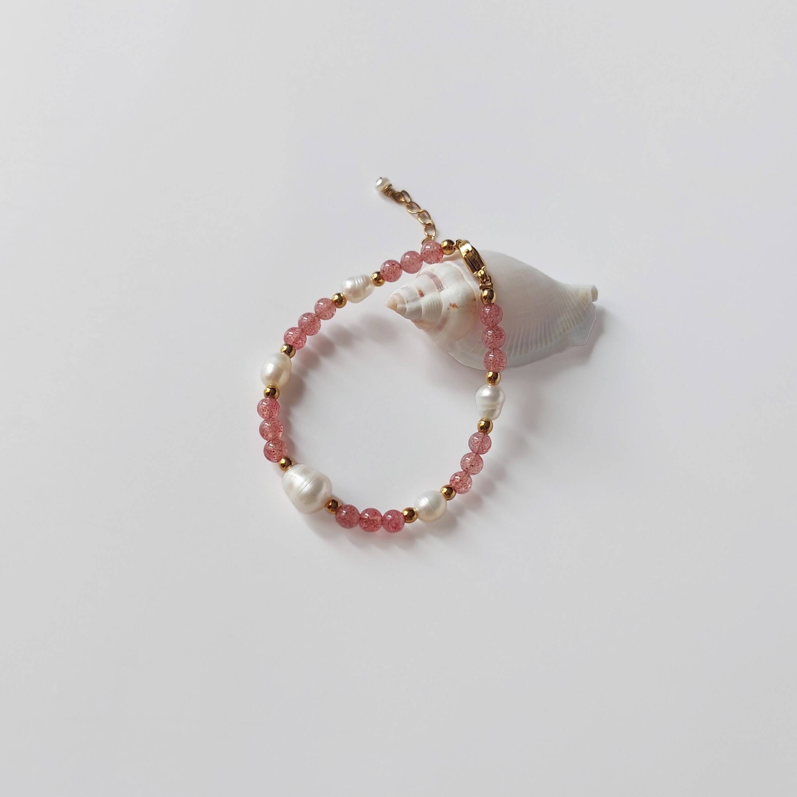 Bracelet Galaxea composé majoritairement de pierres de quartz fraise roses et de perles d'eau douce baroques entrecoupées de perles dorées en acier inoxydable, posé contre un coquillage blanc conique sur fond gris clair.