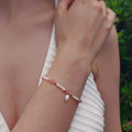 Femme portant le bracelet Millepora en quartz fraise et petites perles d'eau douce baroques entrecoupées de perles dorées en acier inoxydable, photographiée en extérieur avec un haut en crochet blanc et un fond de végétation verte.