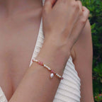 Femme portant le bracelet Millepora en quartz fraise et petites perles d'eau douce baroques entrecoupées de perles dorées en acier inoxydable, photographiée en extérieur avec un haut en crochet blanc et un fond de végétation verte.