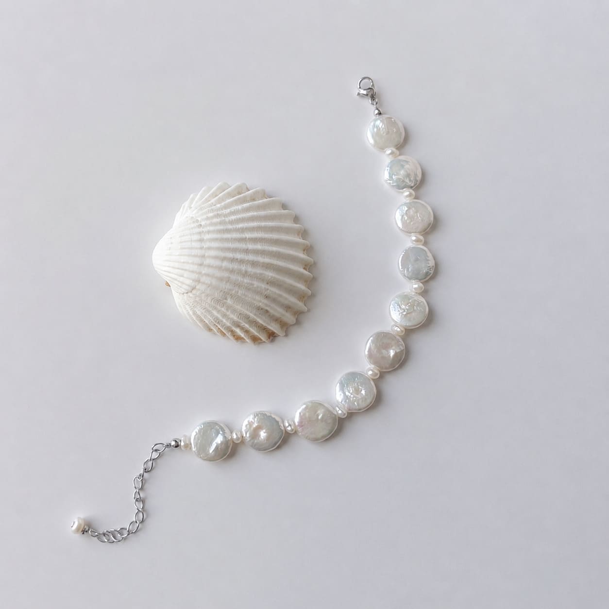 Bracelet en acier inoxydable argenté avec perles d’eau douce naturelles blanches plates, bijou lumineux posé à côté d’un coquillage, sur fond neutre.