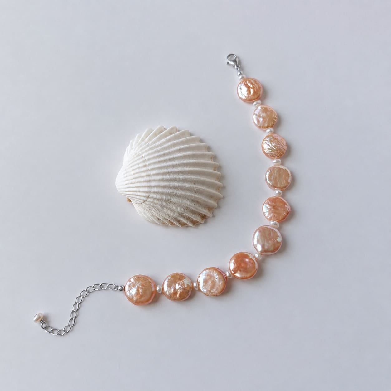 Bracelet en acier inoxydable argenté avec perles d’eau douce naturelles oranges plates, bijou lumineux posé à côté d’un coquillage, sur fond neutre.