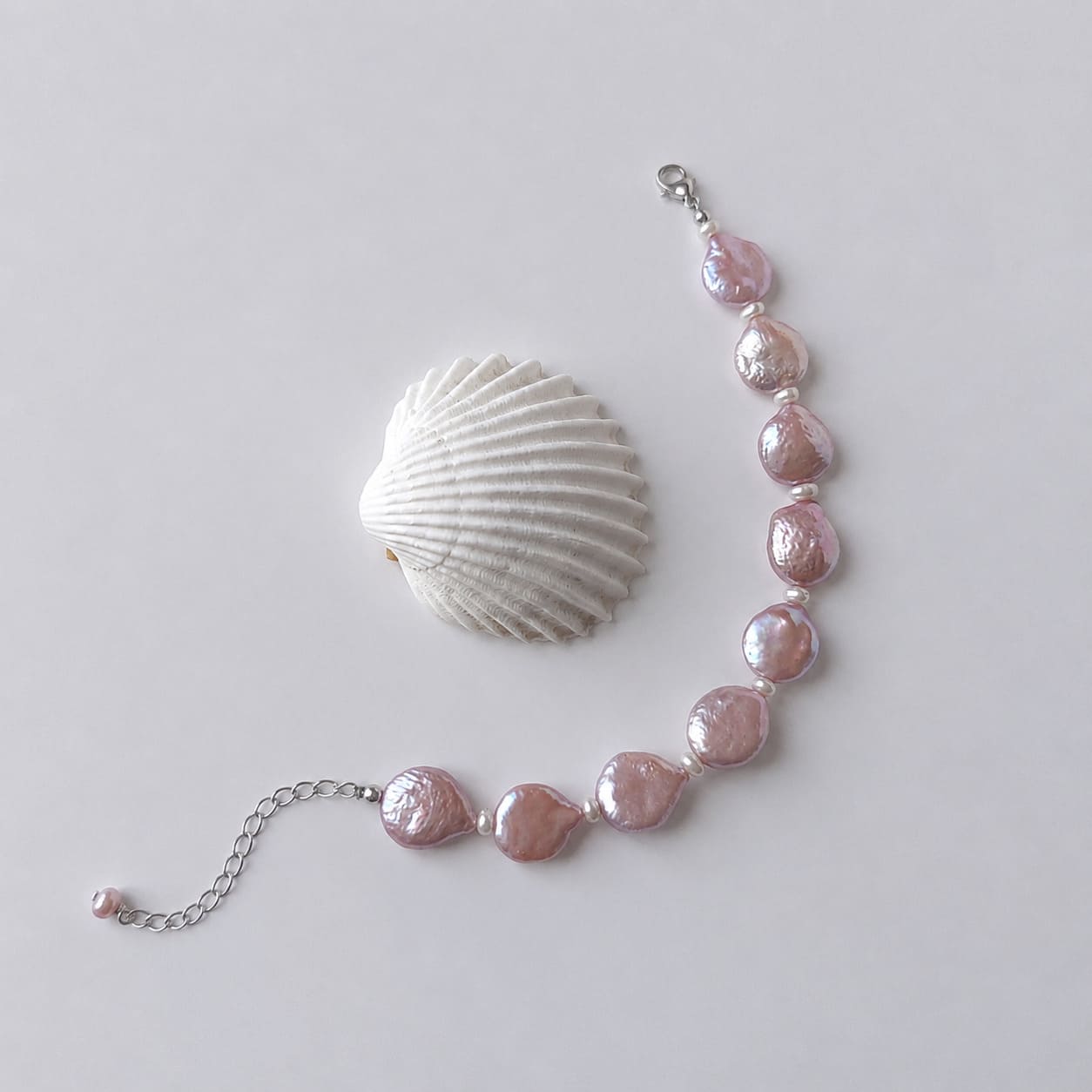 Bracelet en acier inoxydable argenté avec perles d’eau douce naturelles violettes plates, bijou lumineux posé à côté d’un coquillage, sur fond neutre.