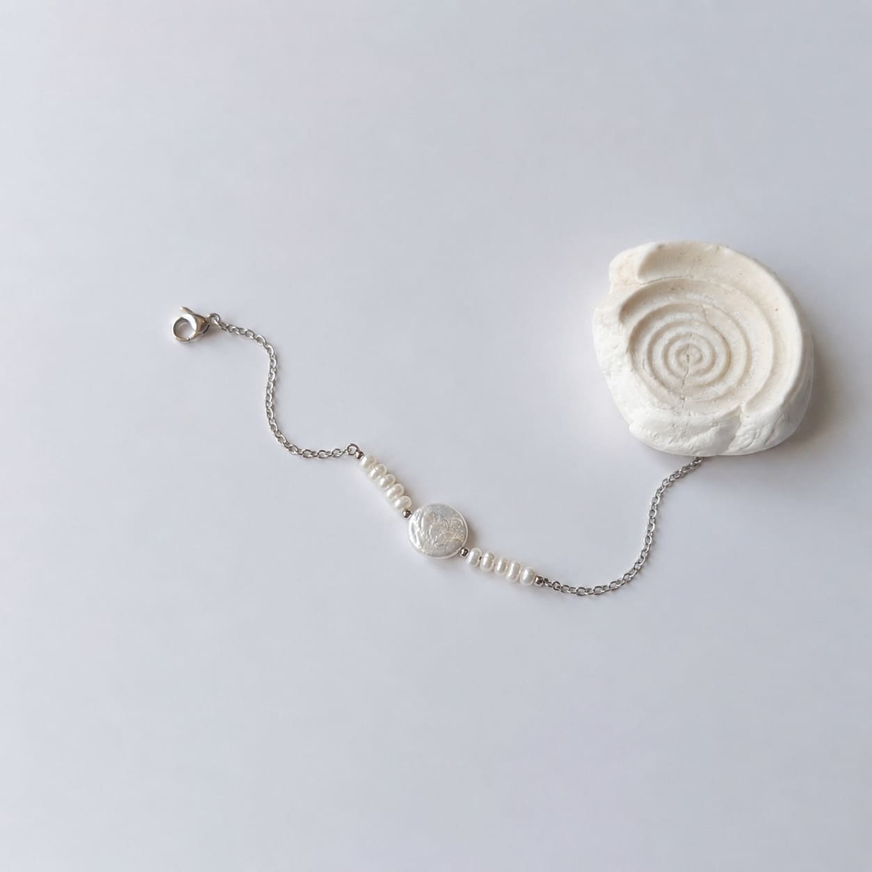 Bracelet Solaris en acier inoxydable argenté avec perles d’eau douce naturelles blanches et perle centrale plate, bijou élégant et minimaliste. Posé à côté d'un coquillage et sur fond neutre.