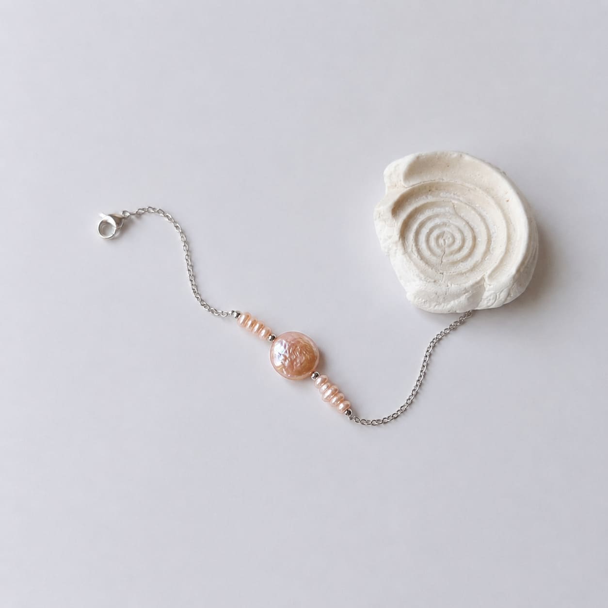 Bracelet Solaris en acier inoxydable argenté avec perles d’eau douce naturelles oranges et perle centrale plate, bijou élégant et minimaliste. Posé à côté d'un coquillage et sur fond neutre.