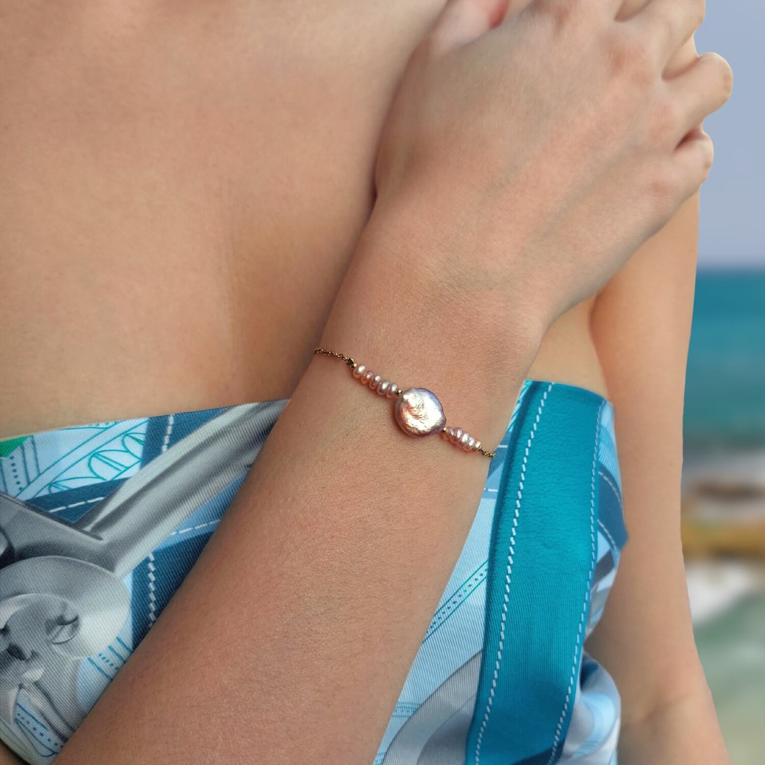 Bracelet en acier inoxydable doré avec perles d’eau douce naturelles violettes et perle centrale plate porté par une femme à la plage