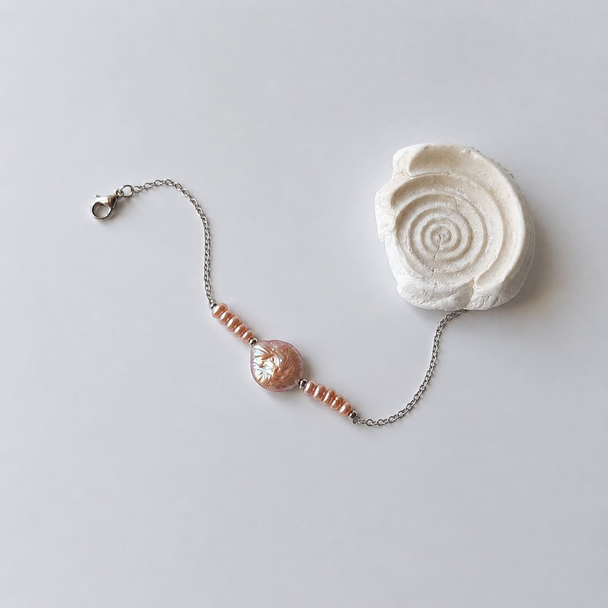 Bracelet Solaris en acier inoxydable argenté avec perles d’eau douce naturelles violettes et perle centrale plate, bijou élégant et minimaliste. Posé à côté d'un coquillage et sur fond neutre.