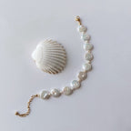 Bracelet en acier inoxydable doré avec perles d’eau douce naturelles blanches plates, bijou lumineux posé à côté d’un coquillage, sur fond neutre.
