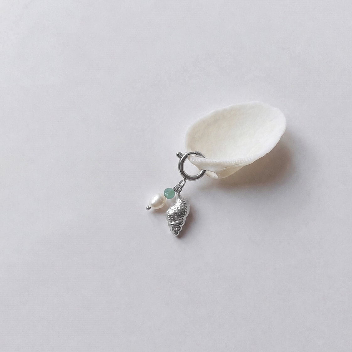 Charm à anneau ressort argenté composé d'une perle d'aventurine, d'une perle d'eau douce blanche et d'un pendentif coquillage conche doré texturé, posé contre un coquillage nacré blanc sur fond gris clair.