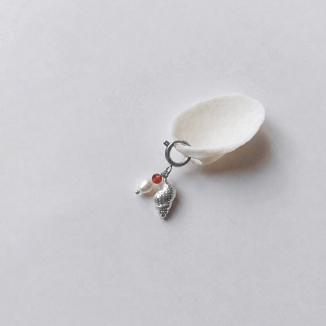 Charm à anneau ressort argenté composé d'une perle de cornaline, d'une perle d'eau douce blanche et d'un pendentif coquillage conche doré texturé, posé contre un coquillage nacré blanc sur fond gris clair.