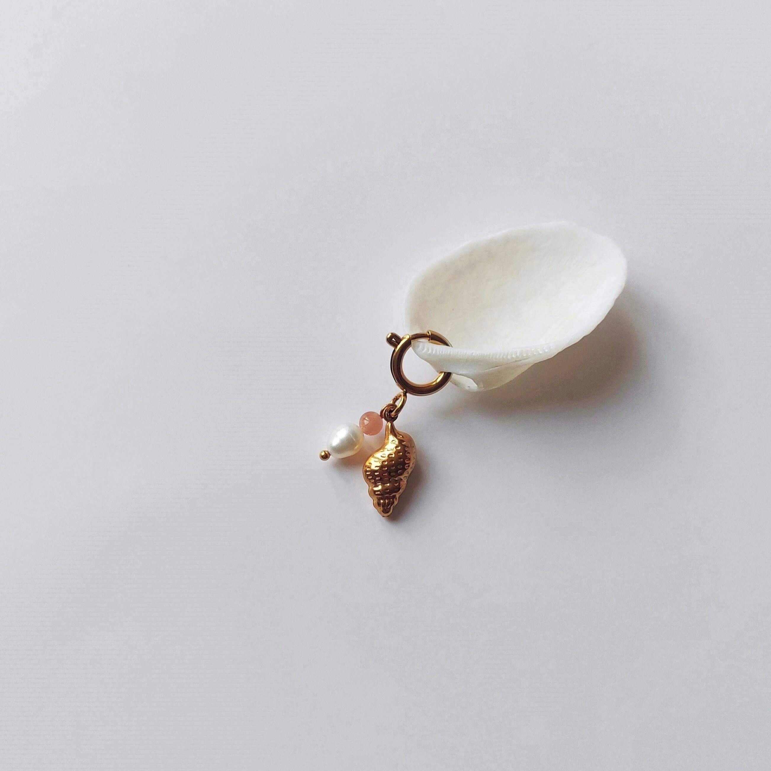 Charm à anneau ressort doré composé d'une pierre de soleil, d'une perle d'eau douce blanche et d'un pendentif coquillage conche doré texturé, posé contre un coquillage nacré blanc sur fond gris clair.