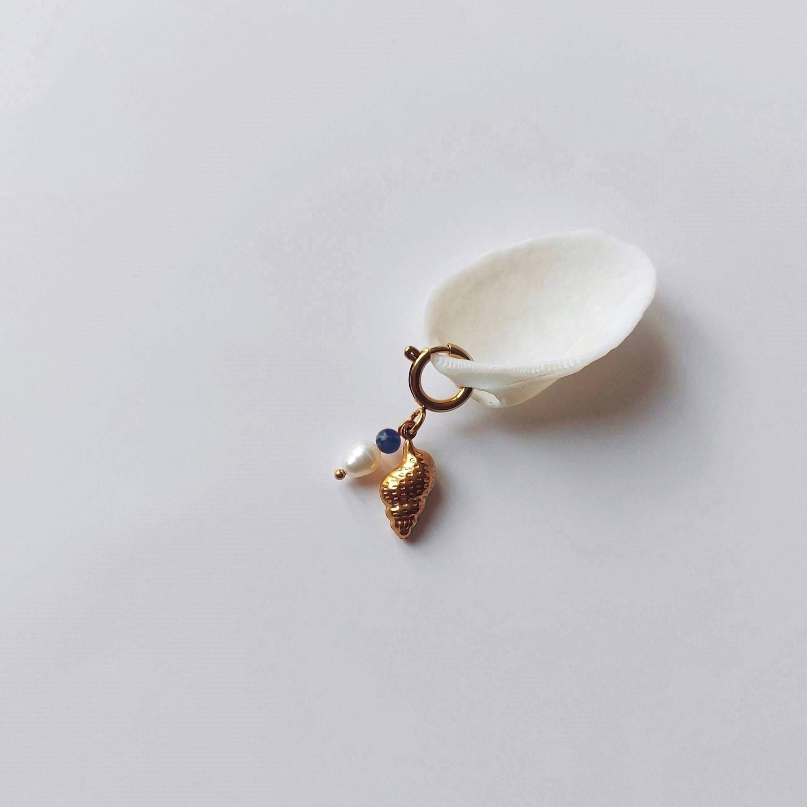 Charm à anneau ressort doré composé d'une pierre de sodalite, d'une perle d'eau douce blanche et d'un pendentif coquillage conche doré texturé, posé contre un coquillage nacré blanc sur fond gris clair.
