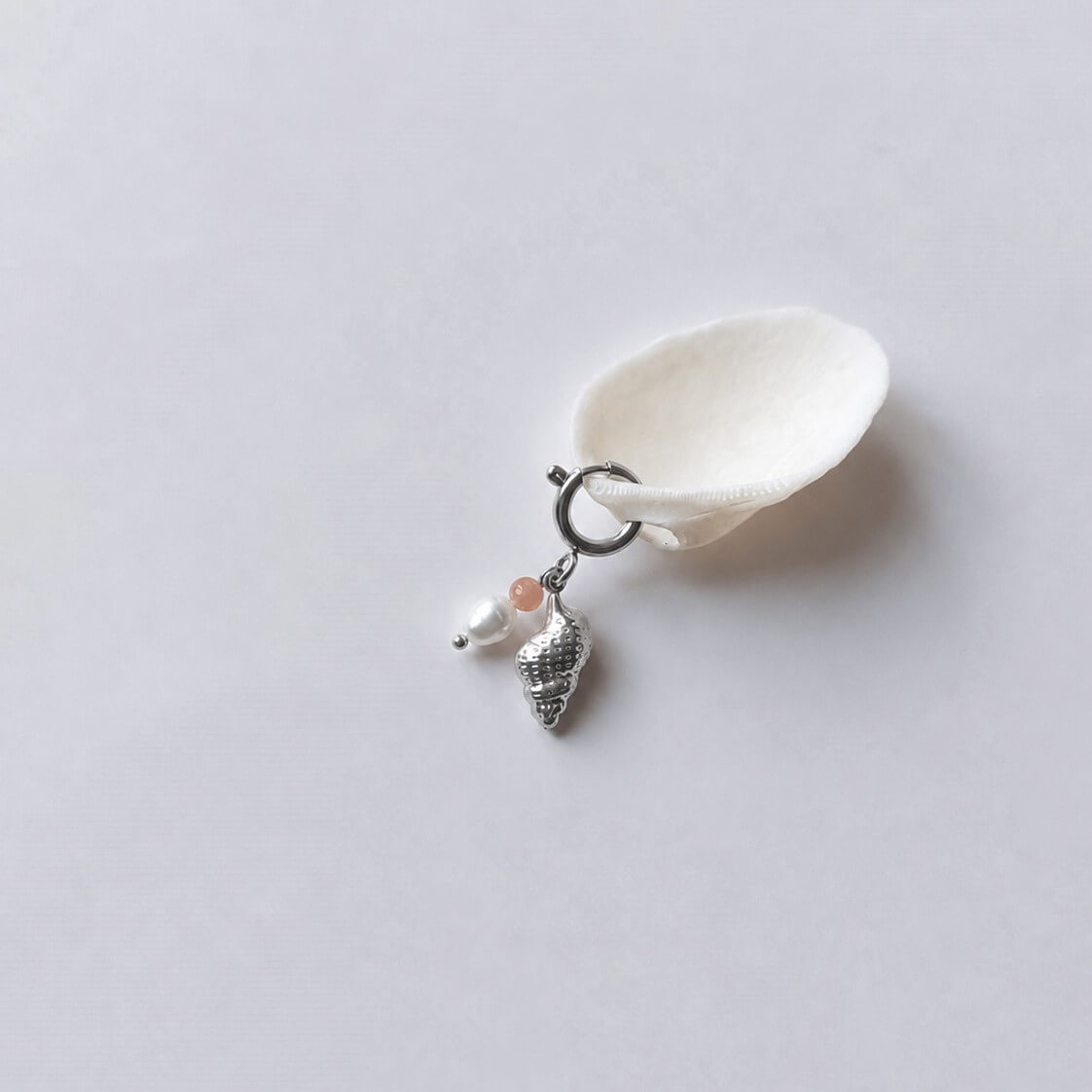 Charm à anneau ressort argenté composé d'une pierre de soleil, d'une perle d'eau douce blanche et d'un pendentif coquillage conche doré texturé, posé contre un coquillage nacré blanc sur fond gris clair.