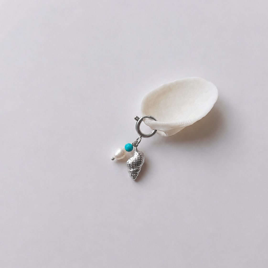 Charm à anneau ressort argenté composé d'une perle de turquoise, d'une perle d'eau douce blanche et d'un pendentif coquillage conche doré texturé, posé contre un coquillage nacré blanc sur fond gris clair.