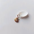 Charm à anneau ressort dorée composé d'une perle d'eau douce blanche, d'une petite perle d'aventurine et d'un pendentif coquille Saint-Jacques dorée striée, posé contre un coquillage nacré blanc sur fond gris clair.