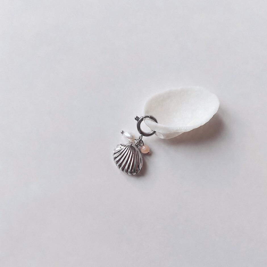 Charm à anneau ressort argenté composé d'une perle d'eau douce blanche, d'une petite perle de pierre de soleil et d'un pendentif coquille Saint-Jacques argentée striée, posé contre un coquillage nacré blanc sur fond gris clair.