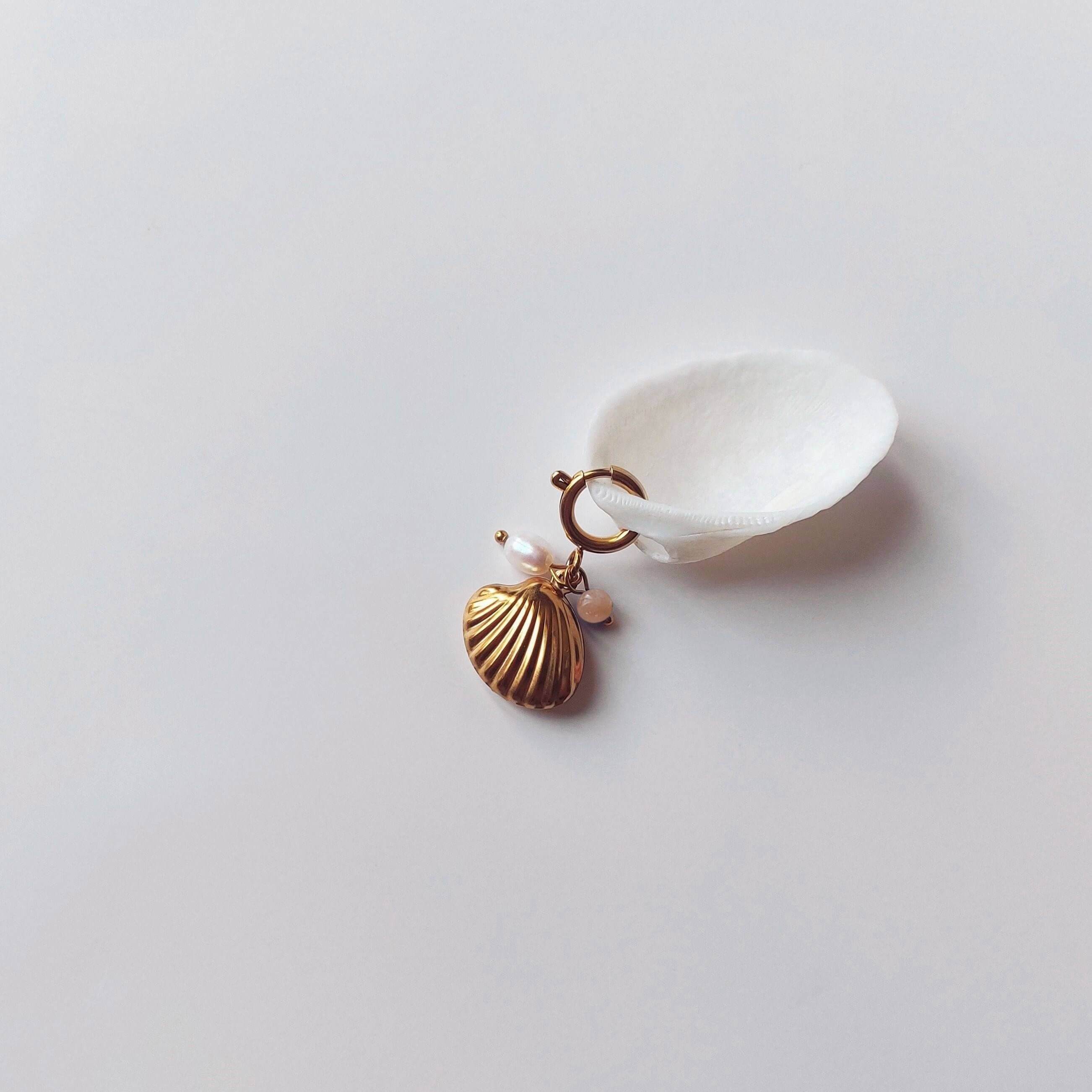 Charm à anneau ressort doré composé d'une perle d'eau douce blanche, d'une petite perle de pierre de soleil et d'un pendentif coquille Saint-Jacques dorée striée, posé contre un coquillage nacré blanc sur fond gris clair.