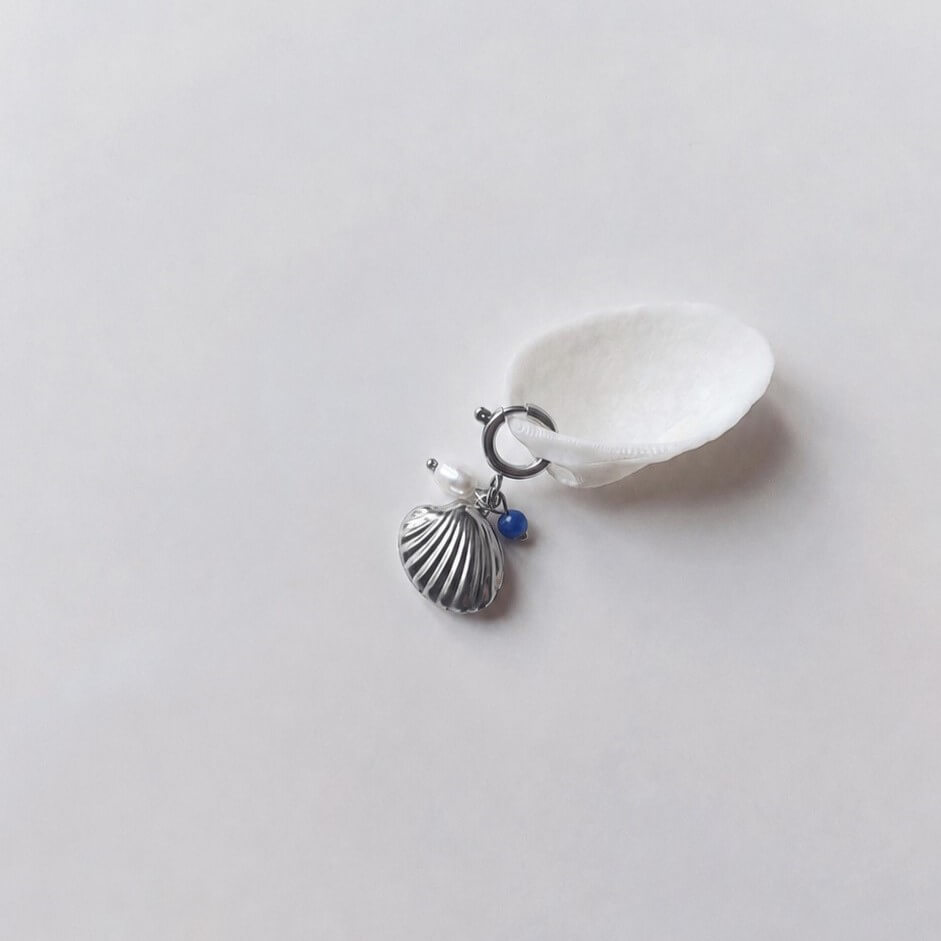 Charm à anneau ressort argenté composé d'une perle d'eau douce blanche, d'une petite perle de sodalite et d'un pendentif coquille Saint-Jacques argentée striée, posé contre un coquillage nacré blanc sur fond gris clair.