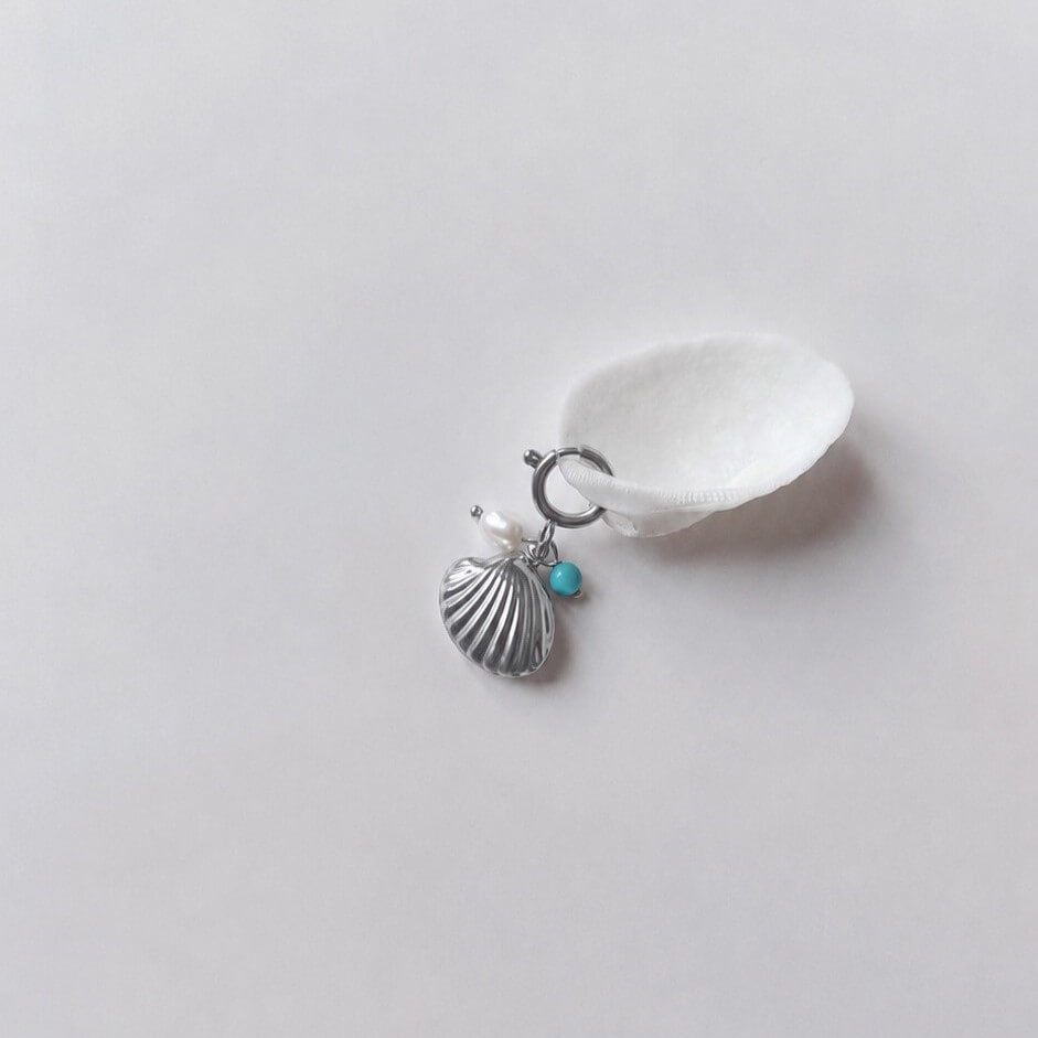 Charm à anneau ressort argenté composé d'une perle d'eau douce blanche, d'une petite perle de turquoise et d'un pendentif coquille Saint-Jacques argentée striée, posé contre un coquillage nacré blanc sur fond gris clair.