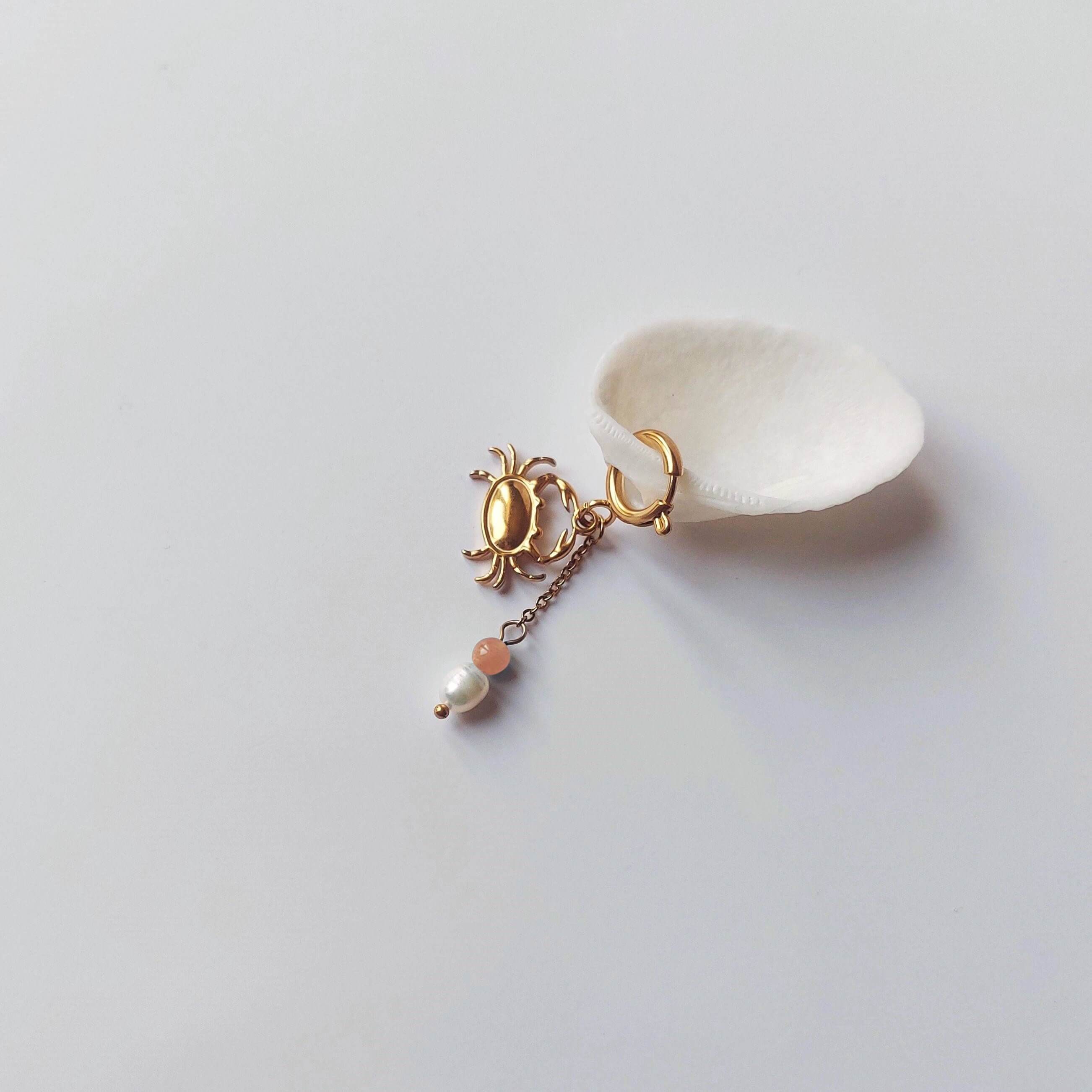 Charm créole dorée en forme de crabe stylisé avec une chaînette fine ornée d'une perle de pierre en pierre de soleil et d'une perle d'eau douce blanche baroque en pendentif, posé contre un coquillage nacré blanc sur fond gris clair.