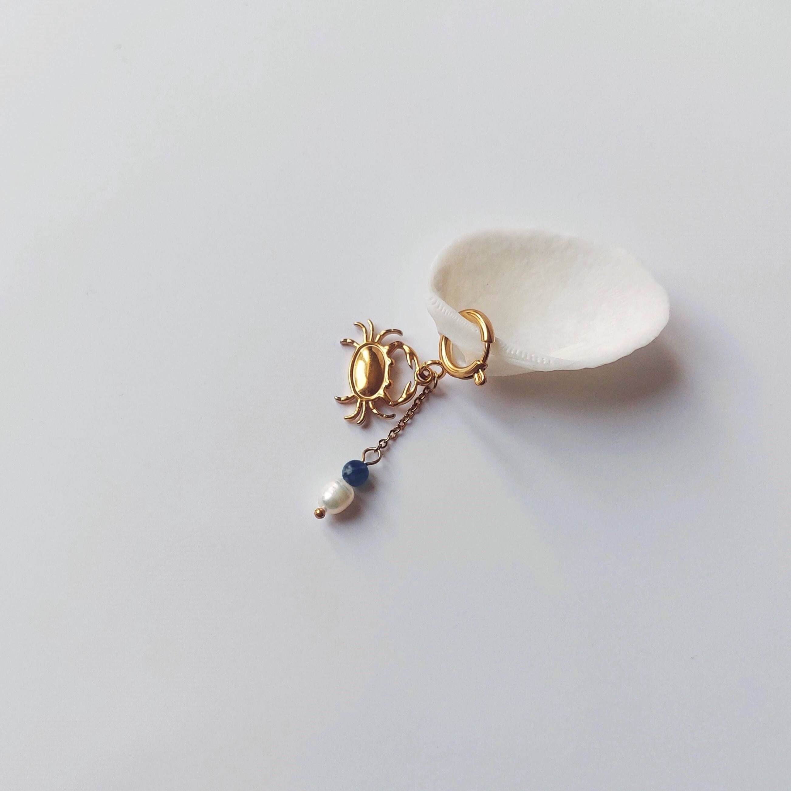 Charm créole dorée en forme de crabe stylisé avec une chaînette fine ornée d'une perle de pierre en sodalite et d'une perle d'eau douce blanche baroque en pendentif, posé contre un coquillage nacré blanc sur fond gris clair.