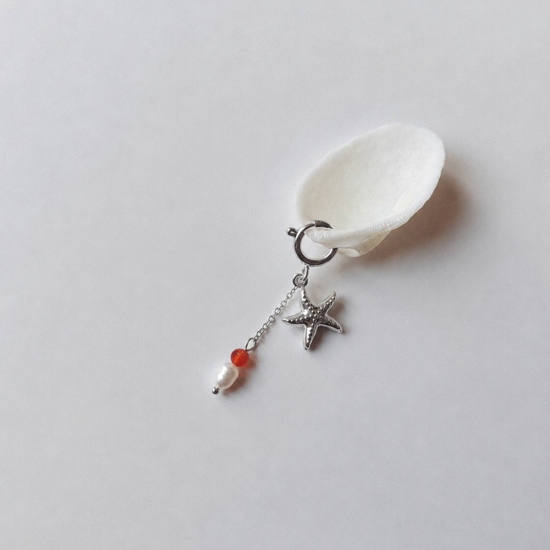 Charm à anneau ressort argenté composé d'un pendentif étoile de mer argentée texturée et d'une chaînette fine ornée d'une petite perle de cornaline et d'une perle d'eau douce blanche baroque, posé contre un coquillage nacré blanc sur fond gris clair.