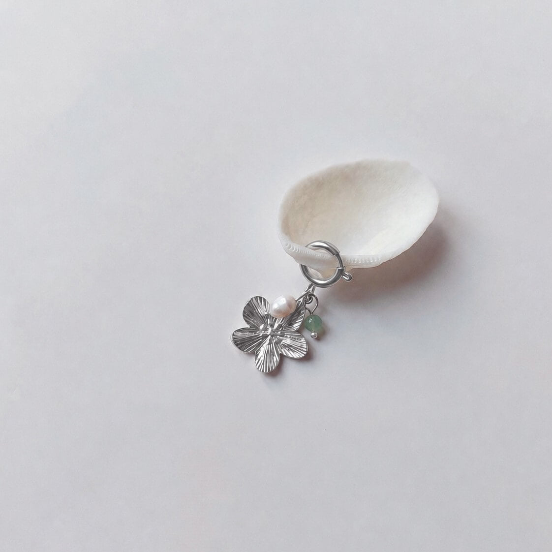 Charm à anneau ressort argenté composé d'une perle d'eau douce blanche, d'une petite perle d'aventurine et d'un pendentif fleur à cinq pétales argentée texturée, posé contre un coquillage nacré blanc sur fond gris clair.
