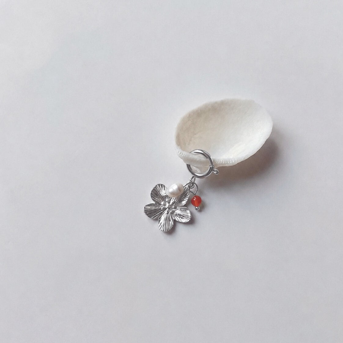 Charm à anneau ressort argenté composé d'une perle d'eau douce blanche, d'une petite perle de cornaline et d'un pendentif fleur à cinq pétales argentée texturée, posé contre un coquillage nacré blanc sur fond gris clair.