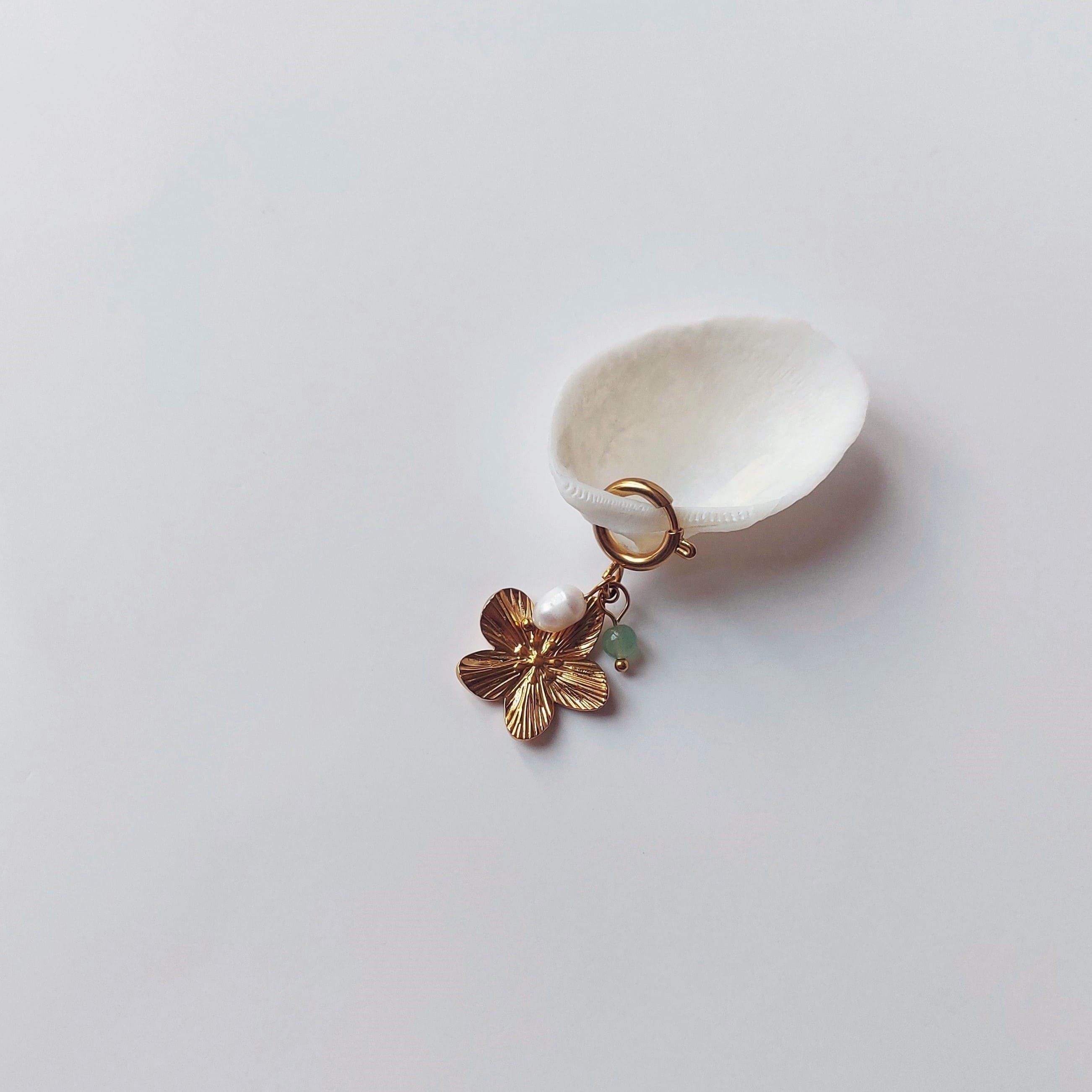 Charm à anneau ressort doré composé d'une perle d'eau douce blanche, d'une petite perle d'aventurine et d'un pendentif fleur à cinq pétales dorée texturée, posé contre un coquillage nacré blanc sur fond gris clair.