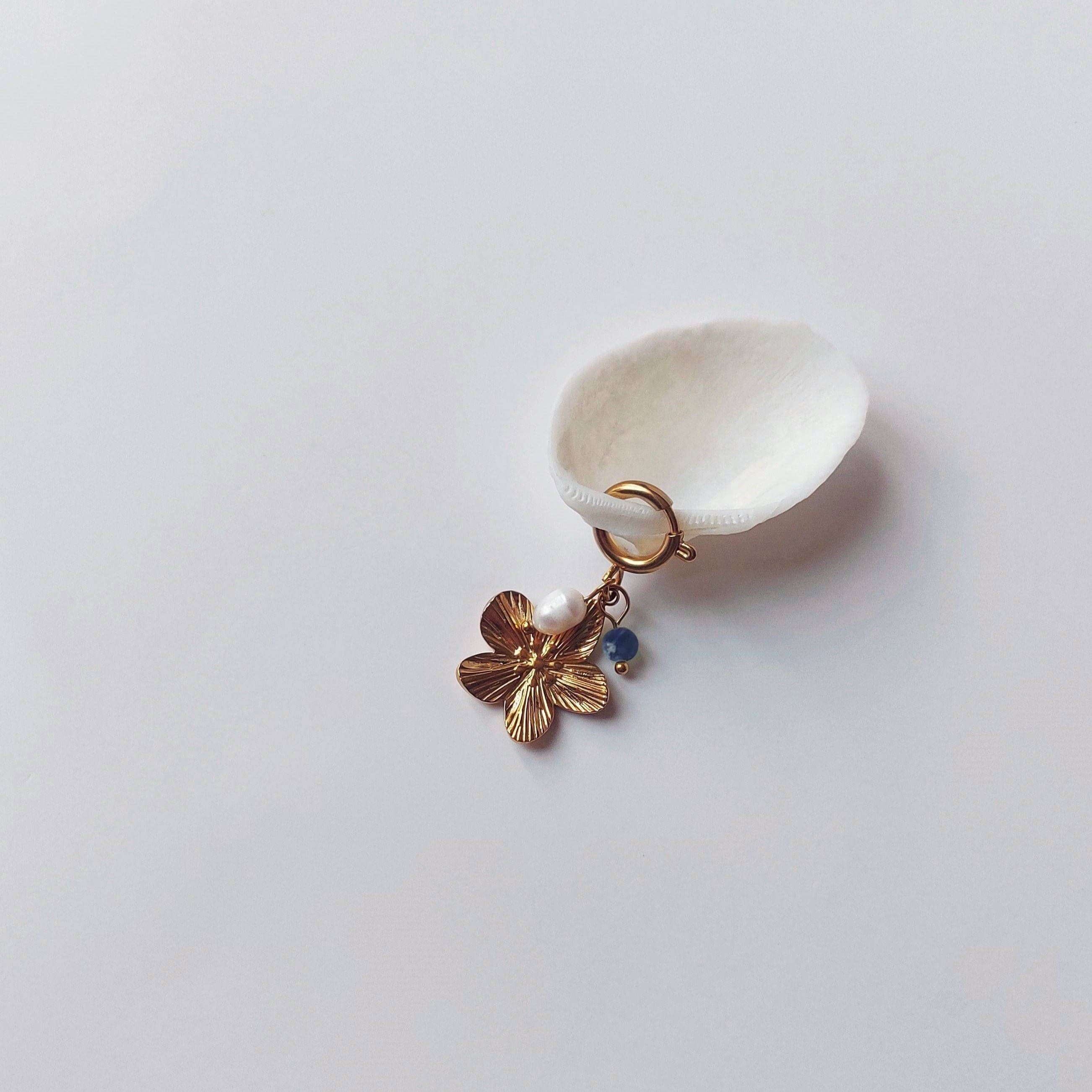 Charm à anneau ressort doré composé d'une perle d'eau douce blanche, d'une petite perle de sodalite et d'un pendentif fleur à cinq pétales dorée texturée, posé contre un coquillage nacré blanc sur fond gris clair.