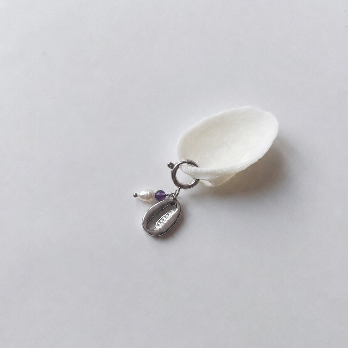Charm à anneau ressort argenté composé d'une perle d'eau douce blanche, d'une petite perle d'améthyste et d'un pendentif cauri argenté, posé contre un coquillage nacré blanc sur fond gris clair.