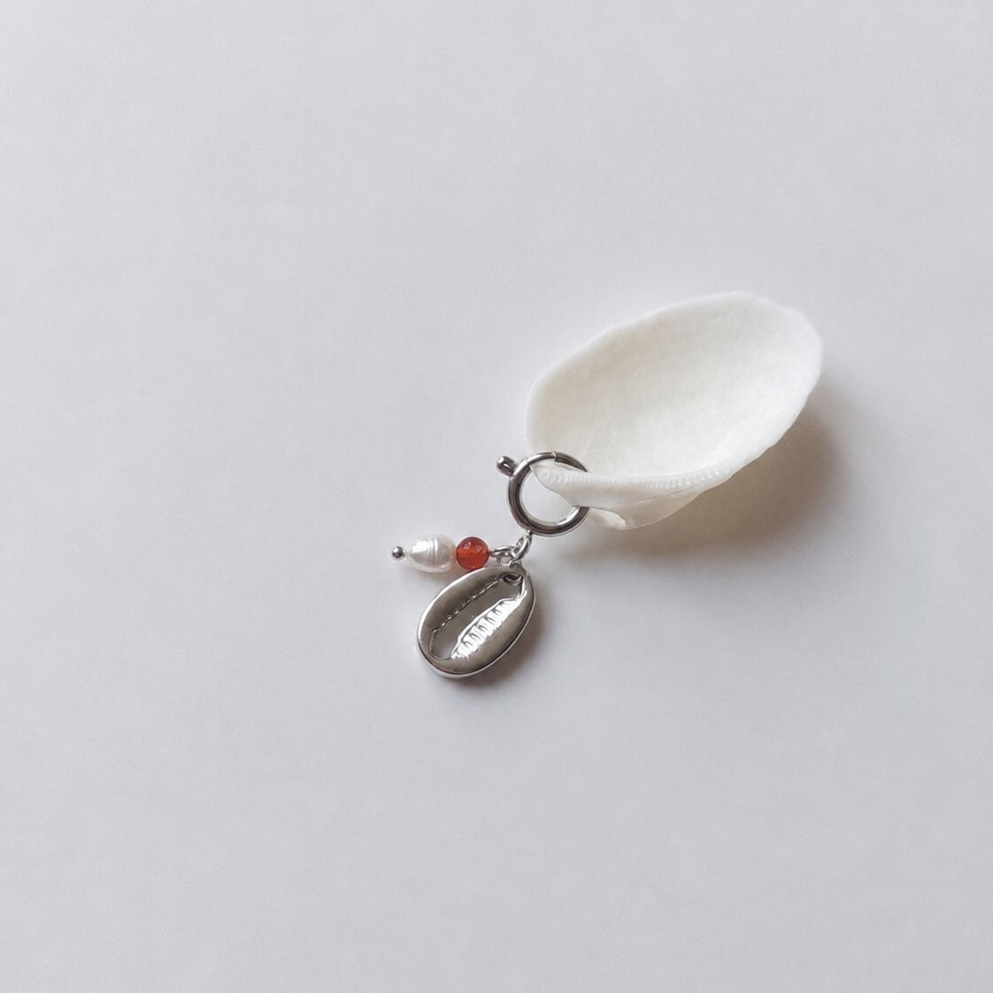 Charm à anneau ressort argenté composé d'une perle d'eau douce blanche, d'une petite perle de cornaline et d'un pendentif cauri argenté, posé contre un coquillage nacré blanc sur fond gris clair.