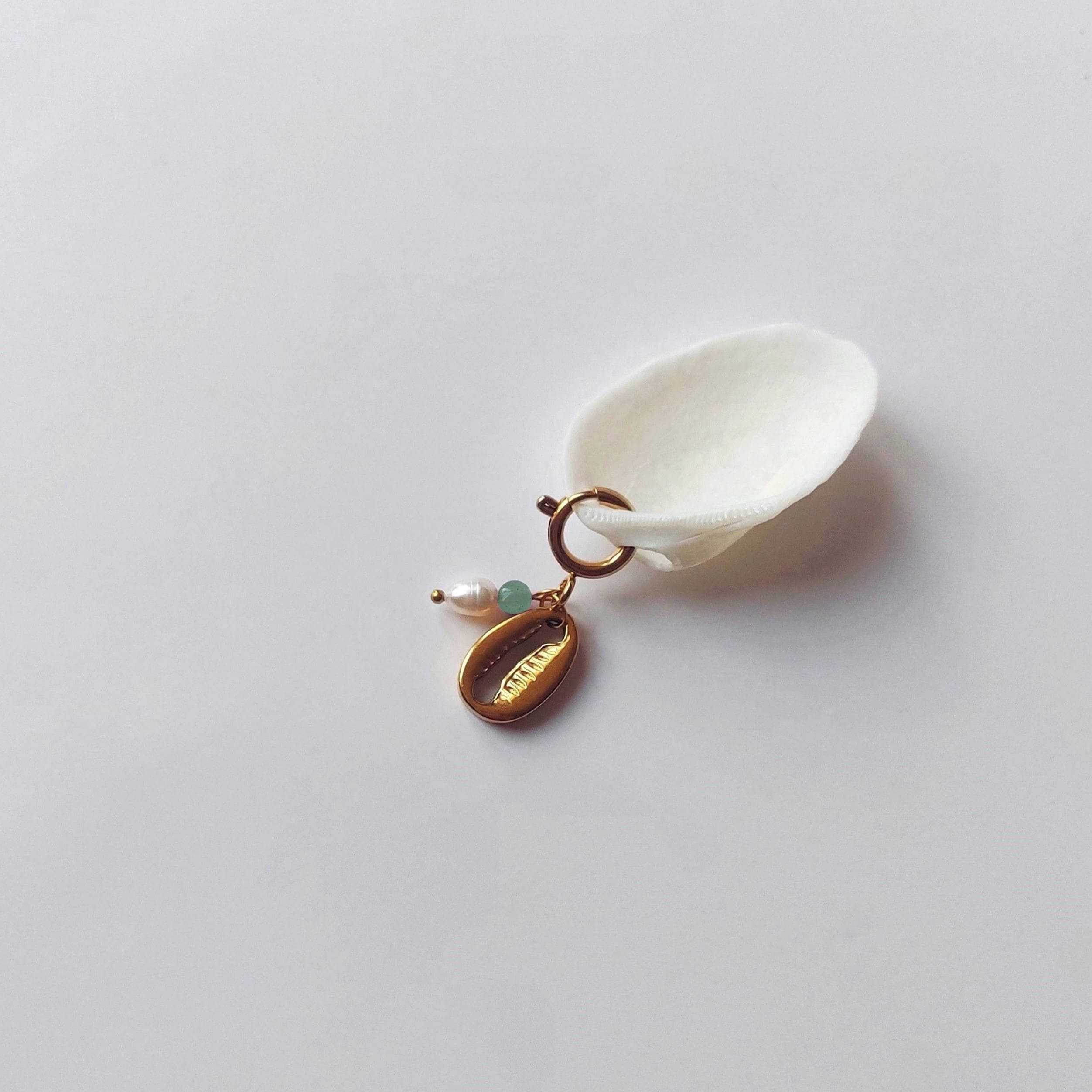 Charm à anneau ressort doré composé d'une perle d'eau douce blanche, d'une petite perle d'aventurine et d'un pendentif cauri doré, posé contre un coquillage nacré blanc sur fond gris clair.
