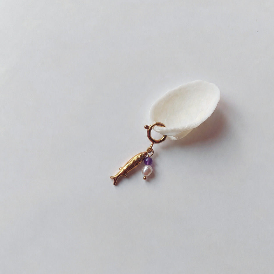 Charm à anneau ressort doré composé d'un pendentif poisson doré gravé, d'une petite perle d'améthyste et d'une perle d'eau douce blanche baroque, posé contre un coquillage nacré blanc sur fond gris clair.