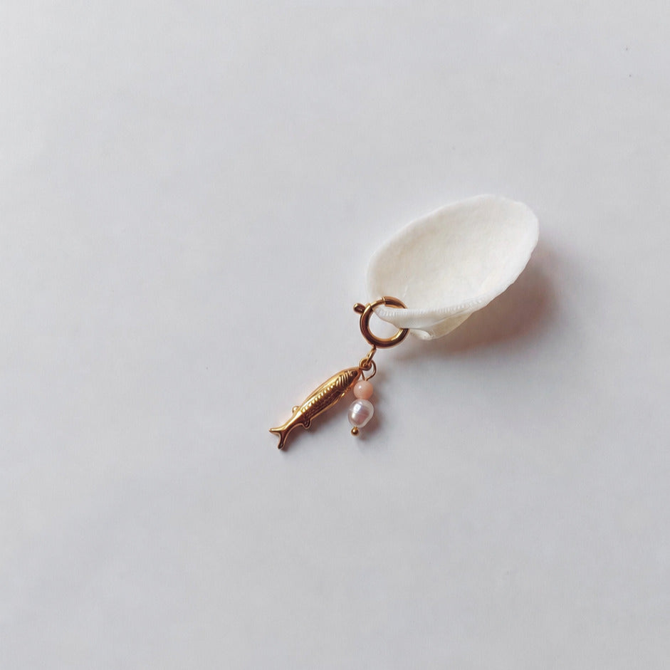 Charm à anneau ressort doré composé d'un pendentif poisson doré gravé, d'une petite perle de pierre de soleil et d'une perle d'eau douce blanche baroque, posé contre un coquillage nacré blanc sur fond gris clair.