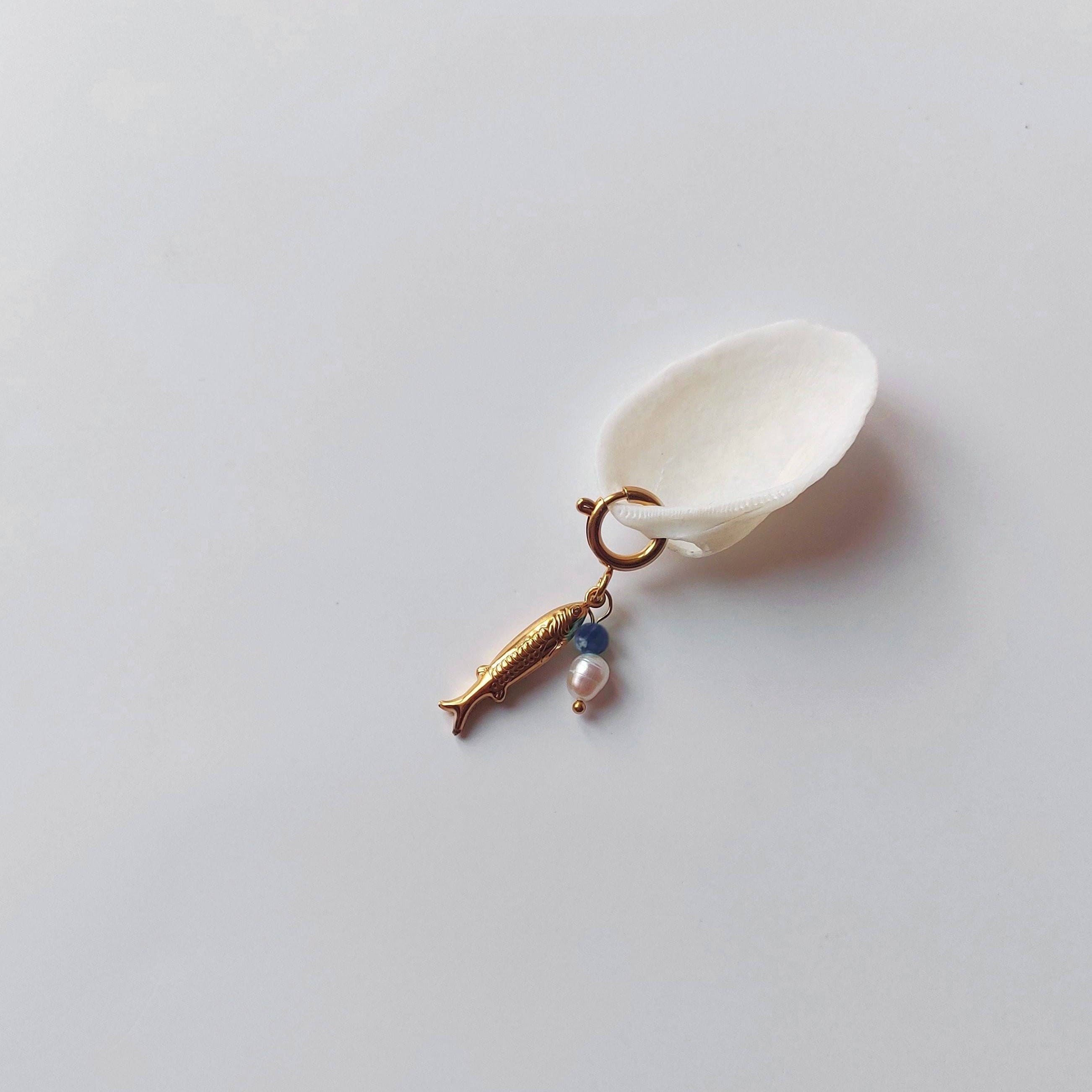 Charm à anneau ressort doré composé d'un pendentif poisson doré gravé, d'une petite perle de sodalite et d'une perle d'eau douce blanche baroque, posé contre un coquillage nacré blanc sur fond gris clair.