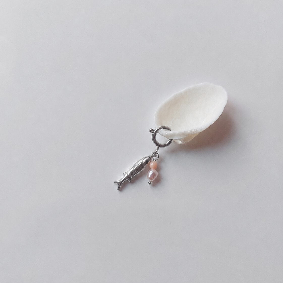 Charm à anneau ressort argenté composé d'un pendentif poisson argenté gravé, d'une petite perle de pierre de soleil et d'une perle d'eau douce blanche baroque, posé contre un coquillage nacré blanc sur fond gris clair.