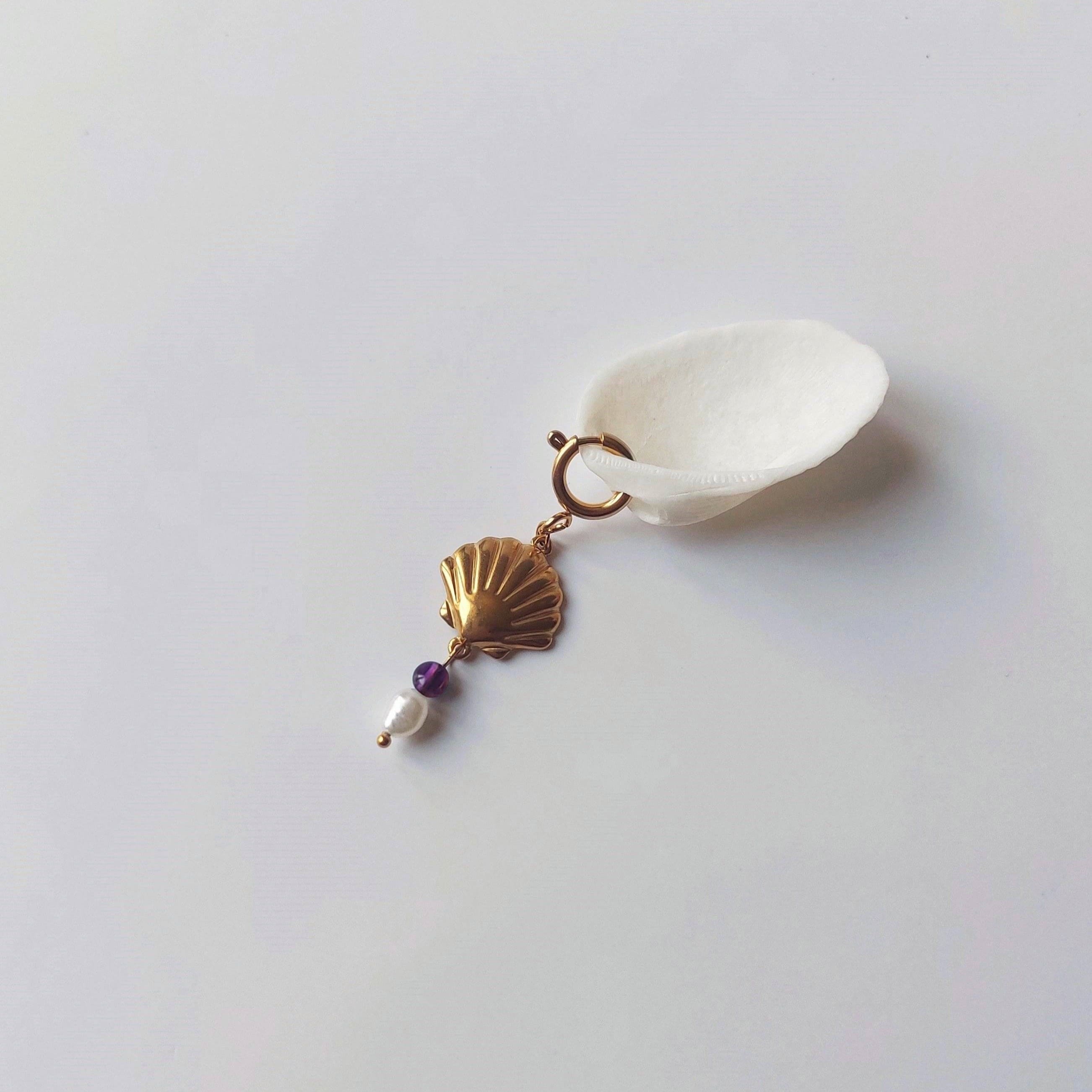 Charm à anneau ressort doré composé d'un pendentif coquille Saint-Jacques dorée striée, d'une petite perle d'améthyste et d'une perle d'eau douce blanche baroque, posé contre un coquillage nacré blanc sur fond gris clair.