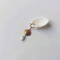 Charm à anneau ressort doré composé d'un pendentif coquille Saint-Jacques dorée striée, d'une petite perle d'aventurine et d'une perle d'eau douce blanche baroque, posé contre un coquillage nacré blanc sur fond gris clair.