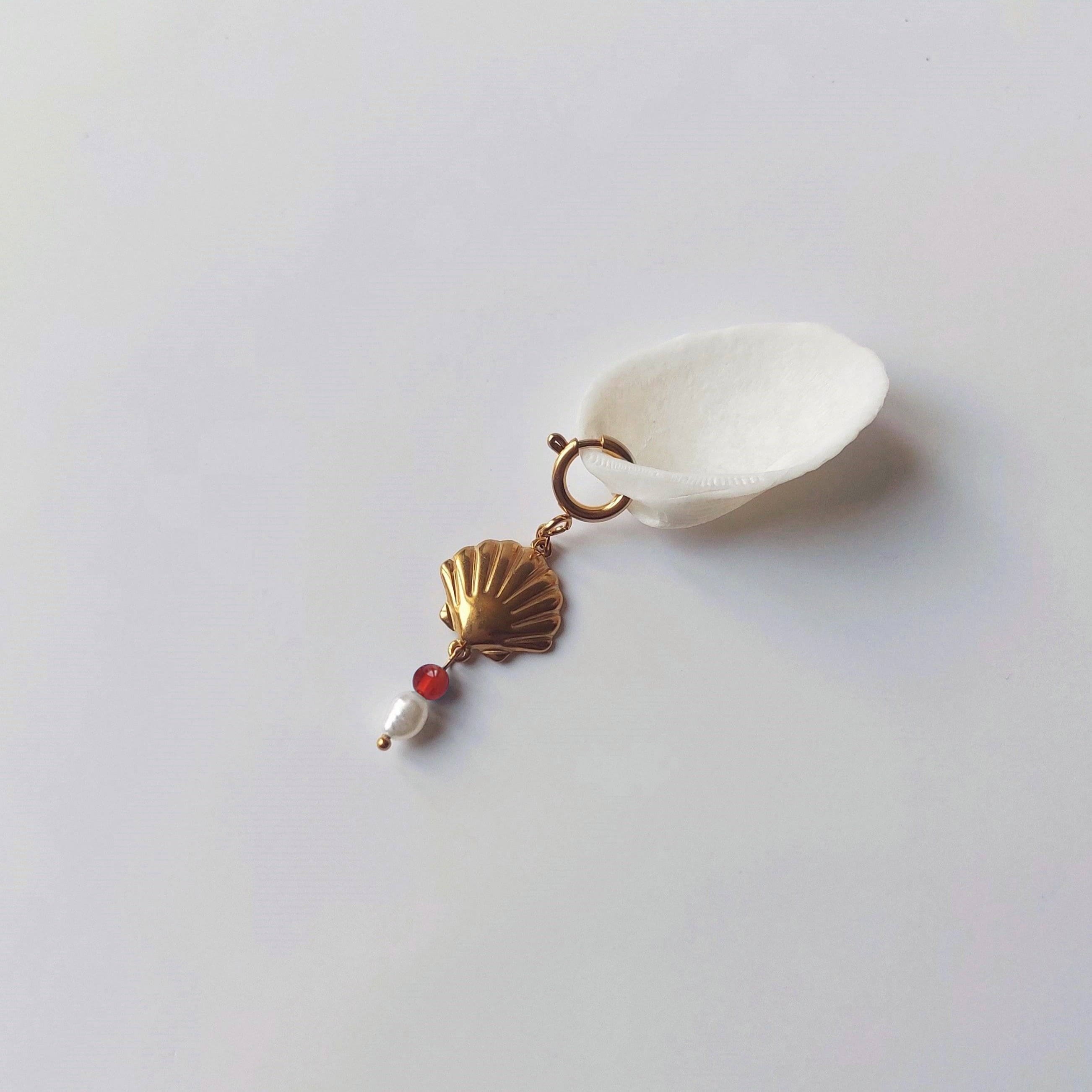 Charm à anneau ressort doré composé d'un pendentif coquille Saint-Jacques dorée striée, d'une petite perle de cornaline et d'une perle d'eau douce blanche baroque, posé contre un coquillage nacré blanc sur fond gris clair.