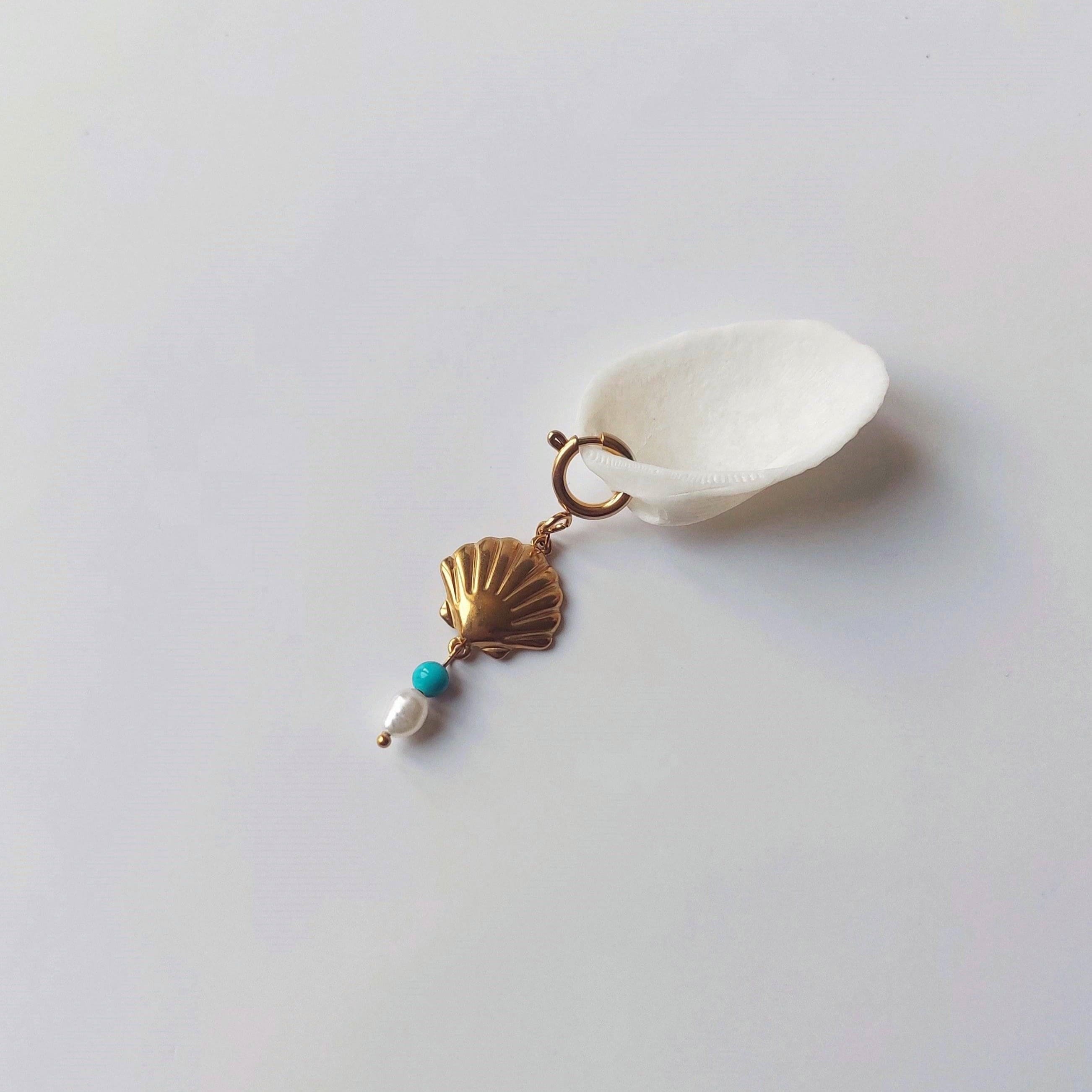 Charm à anneau ressort doré composé d'un pendentif coquille Saint-Jacques dorée striée, d'une petite perle de turquoise bleue et d'une perle d'eau douce blanche baroque, posé contre un coquillage nacré blanc sur fond gris clair.