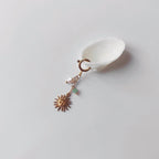 Charm à anneau ressort doré composé d'une perle d'eau douce blanche baroque, d'une chaînette fine ornée d'une petite perle d'aventurine verte et d'un pendentif soleil doré à visage gravé avec rayons, posé contre un coquillage nacré blanc sur fond gris clair.