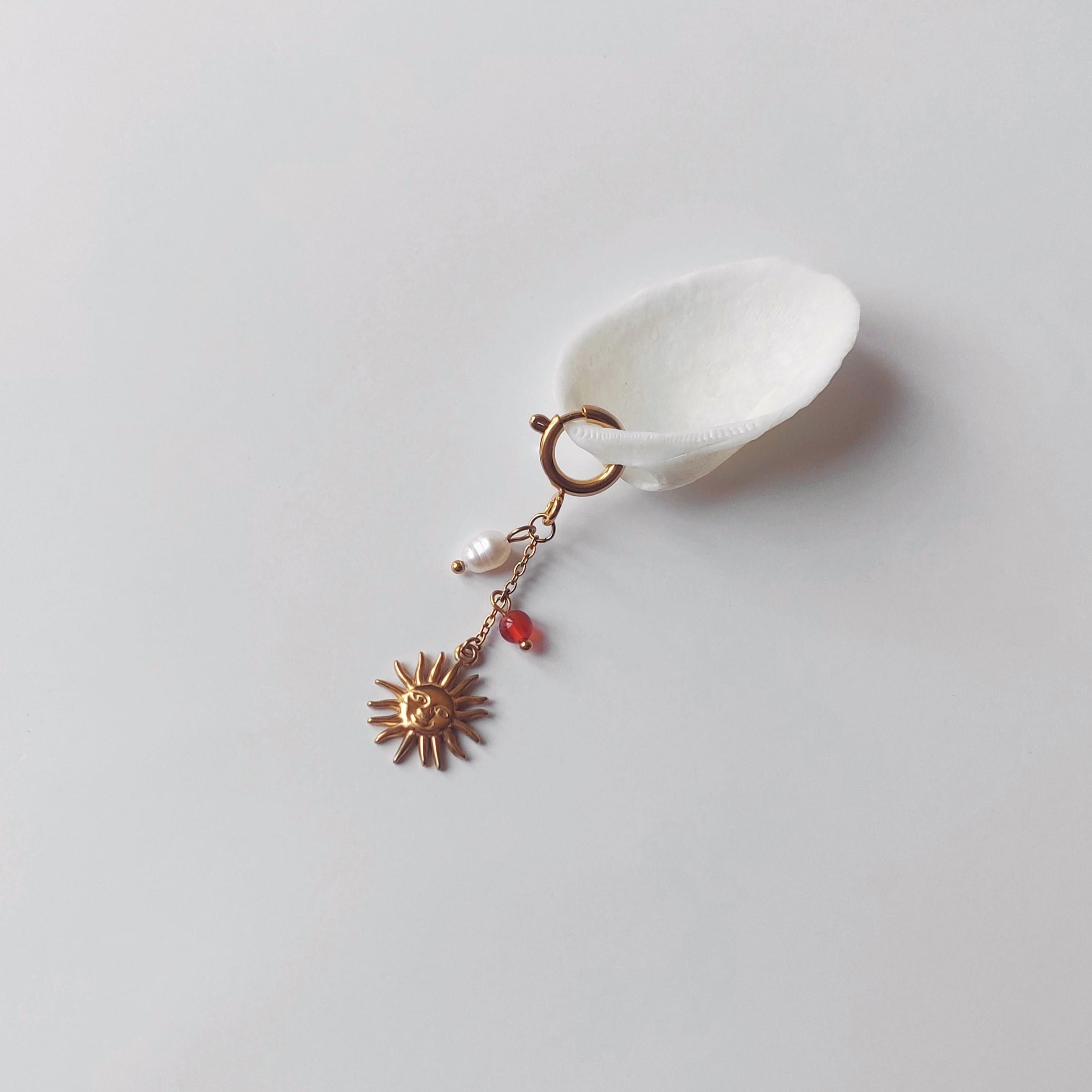 Charm à anneau ressort doré composé d'une perle d'eau douce blanche baroque, d'une chaînette fine ornée d'une petite perle de cornaline et d'un pendentif soleil doré à visage gravé avec rayons, posé contre un coquillage nacré blanc sur fond gris clair.