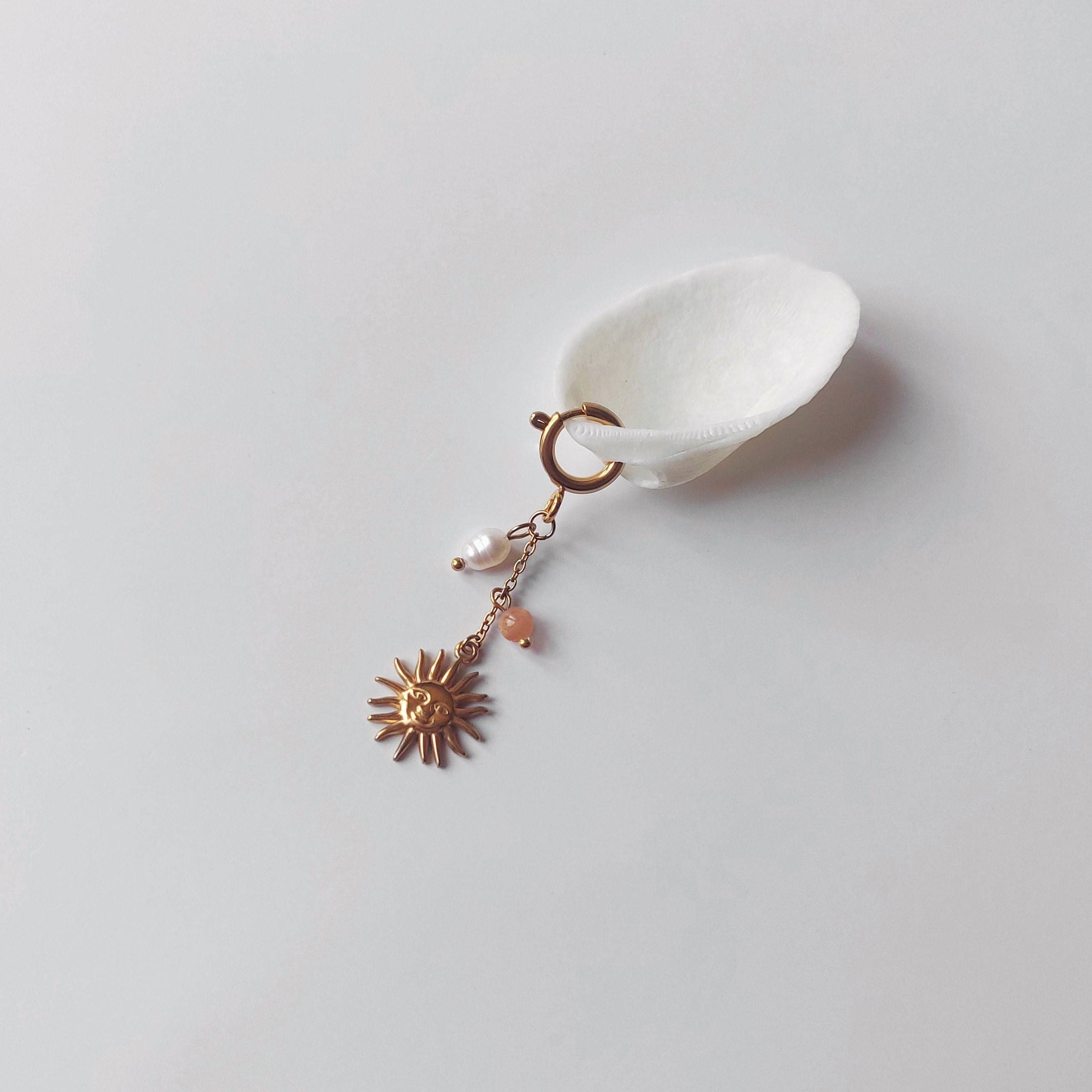 Charm à anneau ressort doré composé d'une perle d'eau douce blanche baroque, d'une chaînette fine ornée d'une petite perle de pierre de soleil et d'un pendentif soleil doré à visage gravé avec rayons, posé contre un coquillage nacré blanc sur fond gris clair.